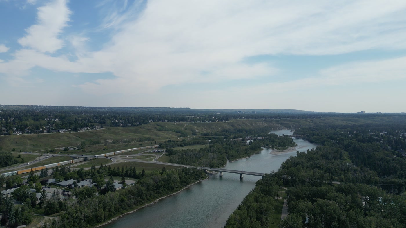 The Bow River at Bowness in Calgary running east under the 85 Street NW bridge, a long freight train on the north bank, truss rail bridges downstream, trees along both banks and high cloud above