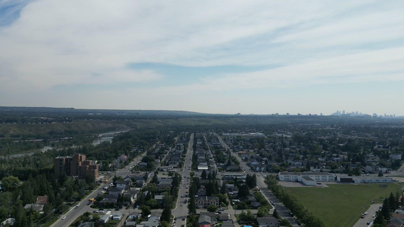 Further east along the avenues of Bowness in Calgary: a brick apartment block close at the left, rows of houses and trees, a long white school with its playing field, downtown's towers on the horizon