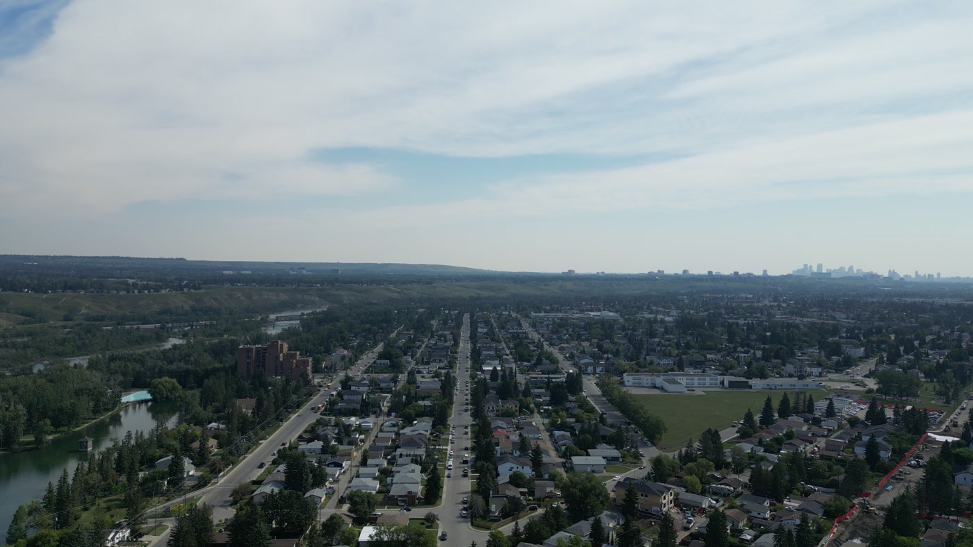 Bowness in Calgary with Bowness Park's lagoon at the lower left: the turquoise wading pool at its end, a brick apartment block, parallel avenues running toward downtown's towers and a long school beside its field