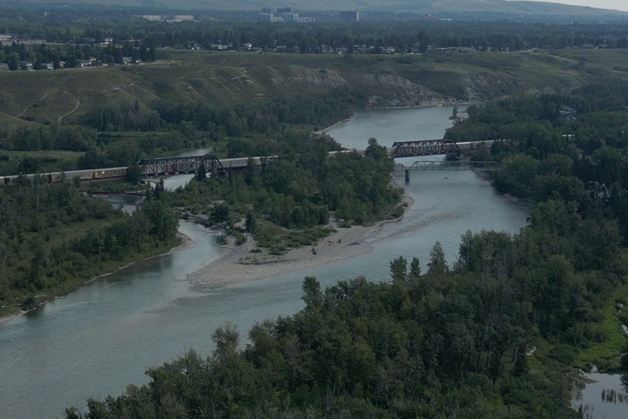A long freight train crossing the Bow River at Bowness in Calgary on two steel truss spans, a lower bridge beneath the second, a pale gravel bar in the current and grassy slopes behind
