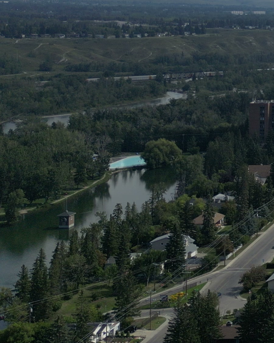 Bowness Park's lagoon in Calgary, a small square pavilion standing in the still water, the turquoise wading pool at its far end, a shore path, houses and a street at the right, the Bow beyond