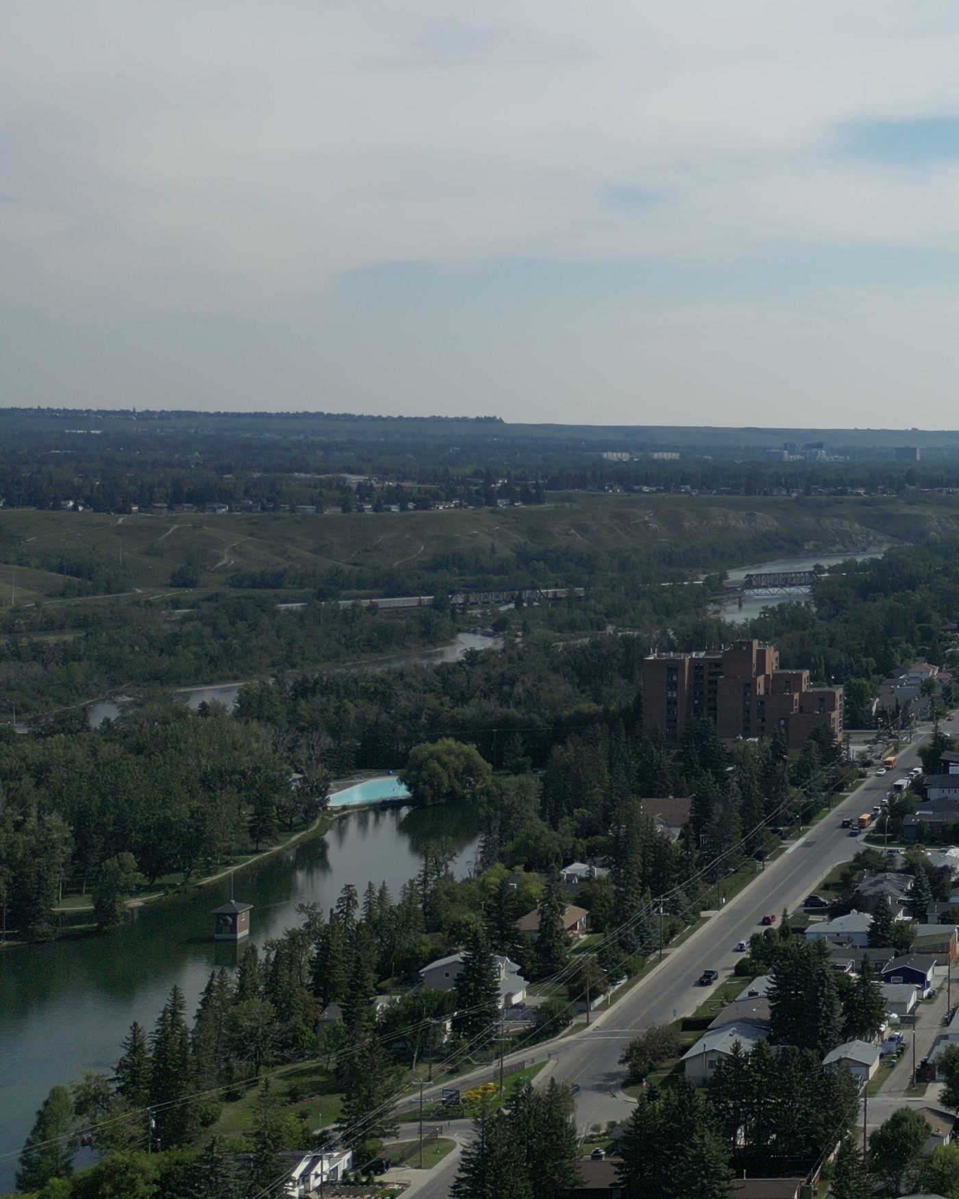 The corner of Bowness Park's lagoon in Calgary: the turquoise wading pool at its end, a pavilion standing in the water, a brick apartment block, a busy avenue, the Bow and its escarpment beyond