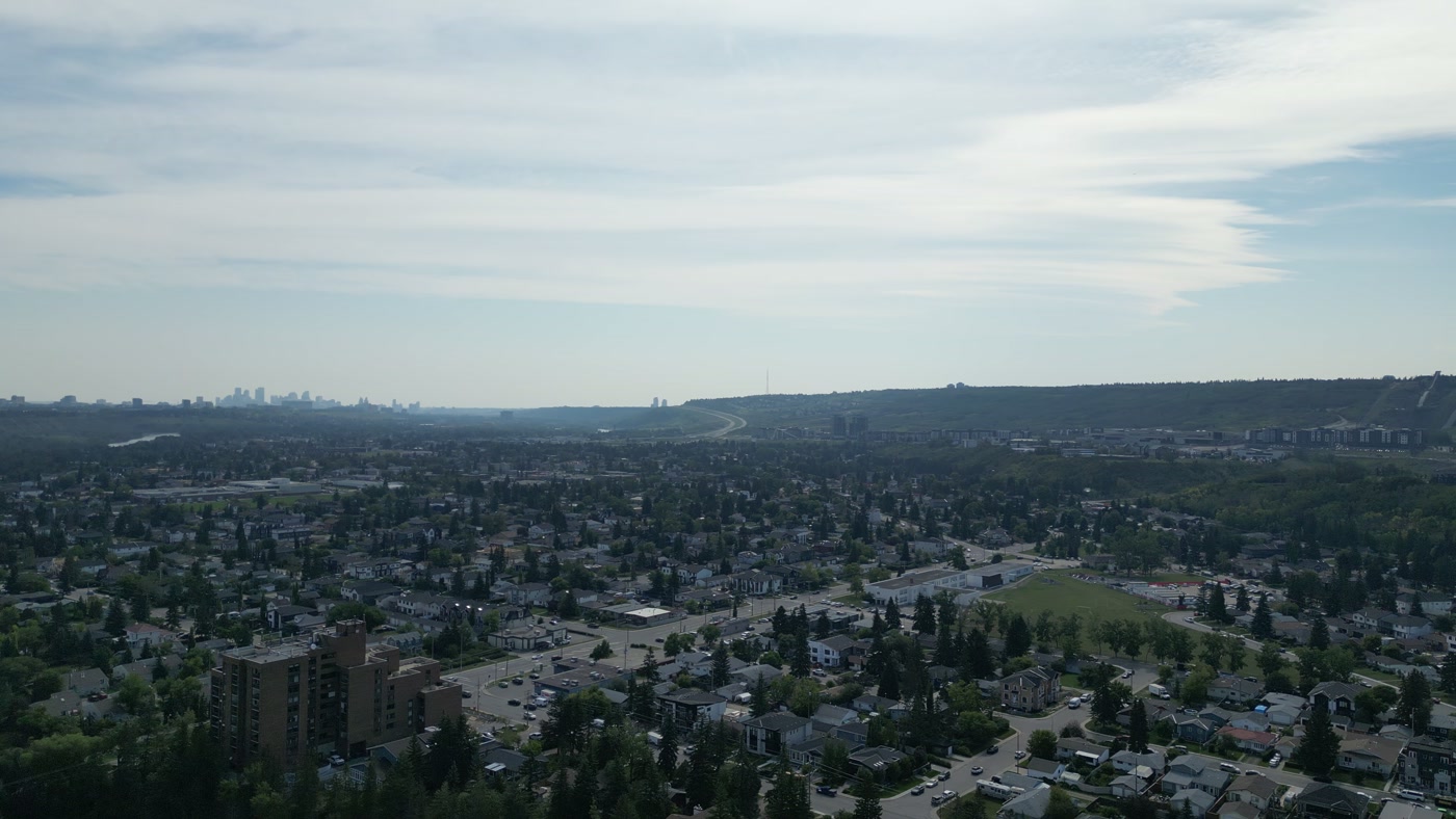 The roofs and trees of Bowness in Calgary, a brick apartment block in front, a school field, the highway climbing a green hill with a ski-jump in-run, and downtown's towers far to the left