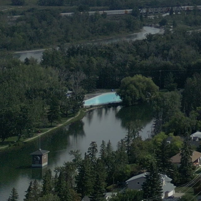 The turquoise wading pool at the east end of Bowness Park's lagoon in Calgary, seen close in the same frame, the lagoon's pavilion below it, trees crowding the bank and the Bow beyond