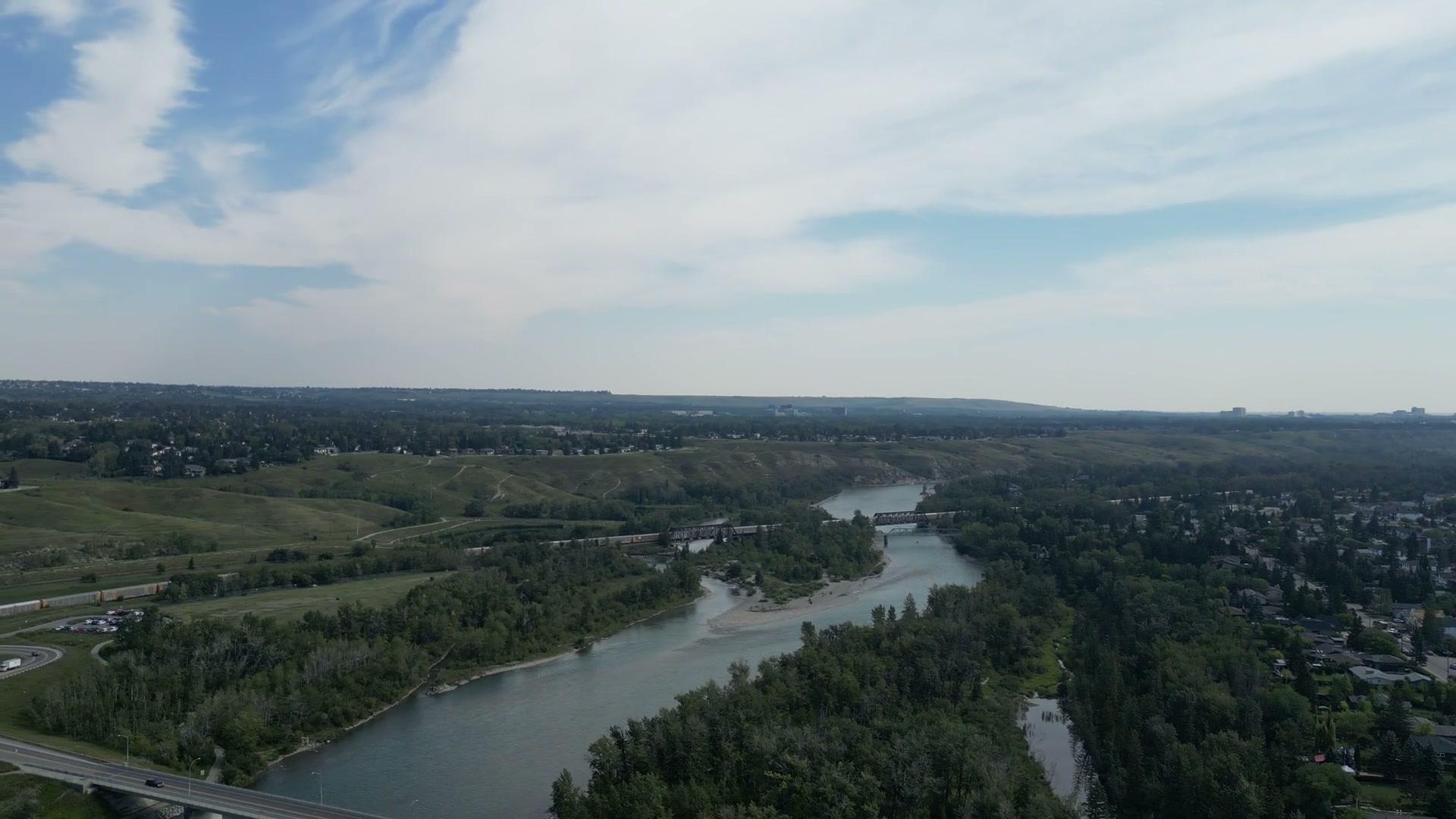 Panning left at half speed from the roofs of Bowness in Calgary back to the Bow, its gravel islands and side channels, a rail line crossing on truss spans and grassy slopes beyond