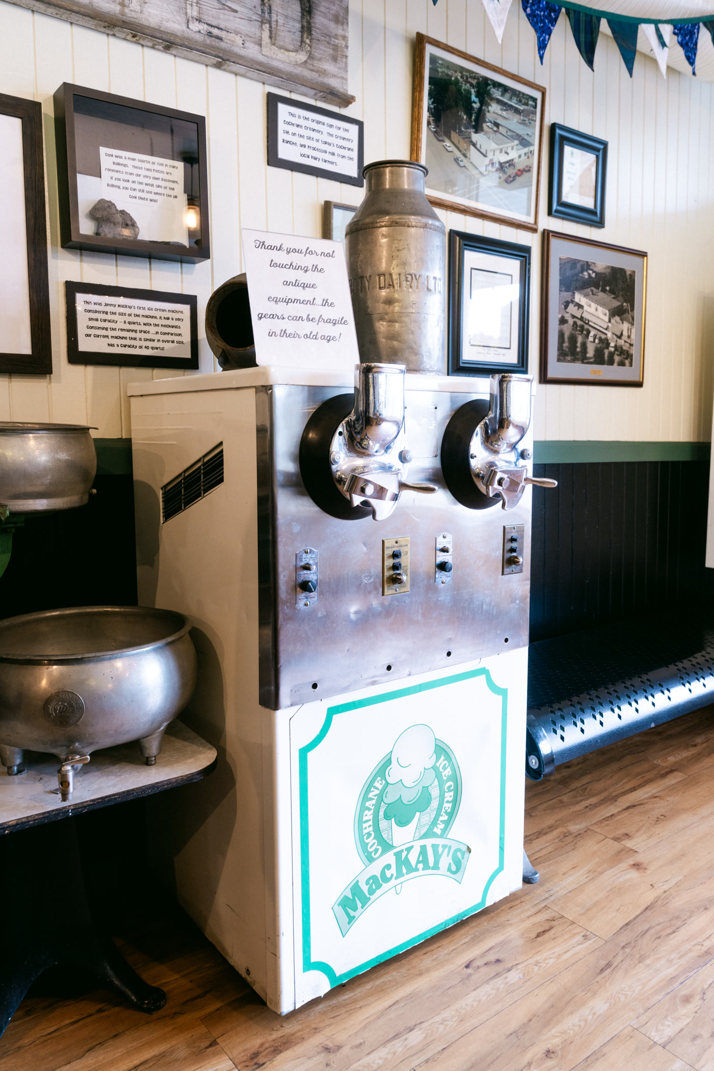 An old stainless ice-cream machine on the shop floor, two spouts over a steel bowl, the MacKay's roundel on the panel below, framed photographs behind it