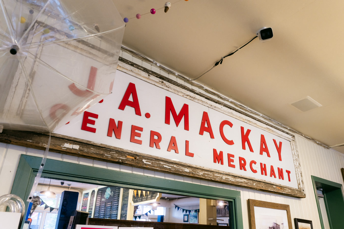 An older J. A. MacKay General Merchant sign, red block letters on a weathered white board, hung high on the shop wall above a green-trimmed doorway