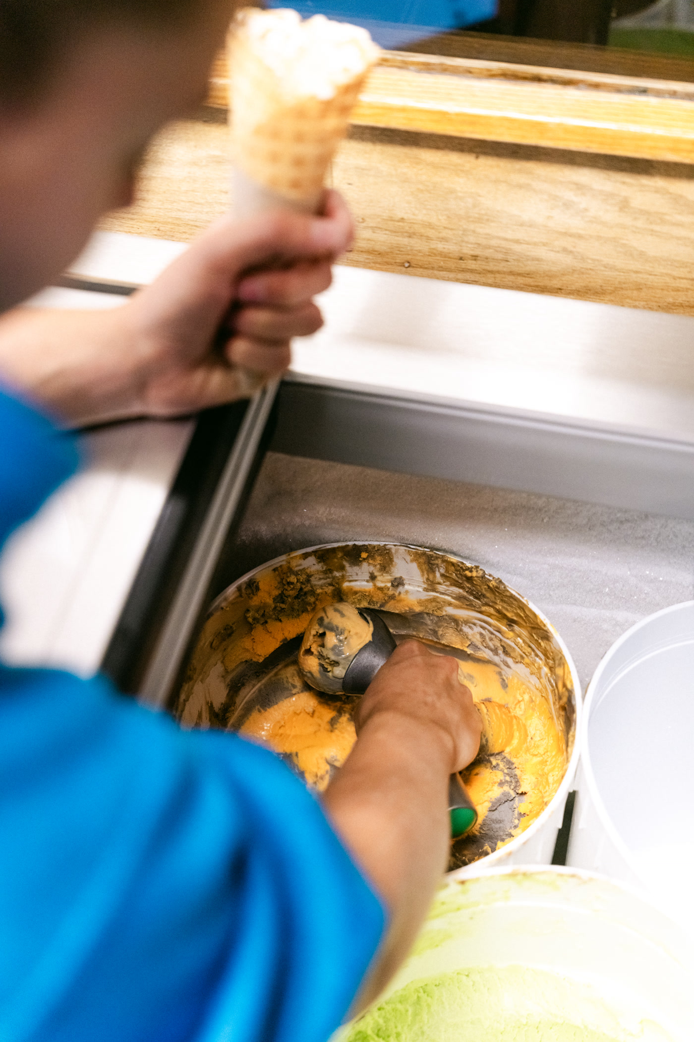 A hand in a blue sleeve scooping caramel-coloured ice cream from a tub into a waffle cone held over the freezer