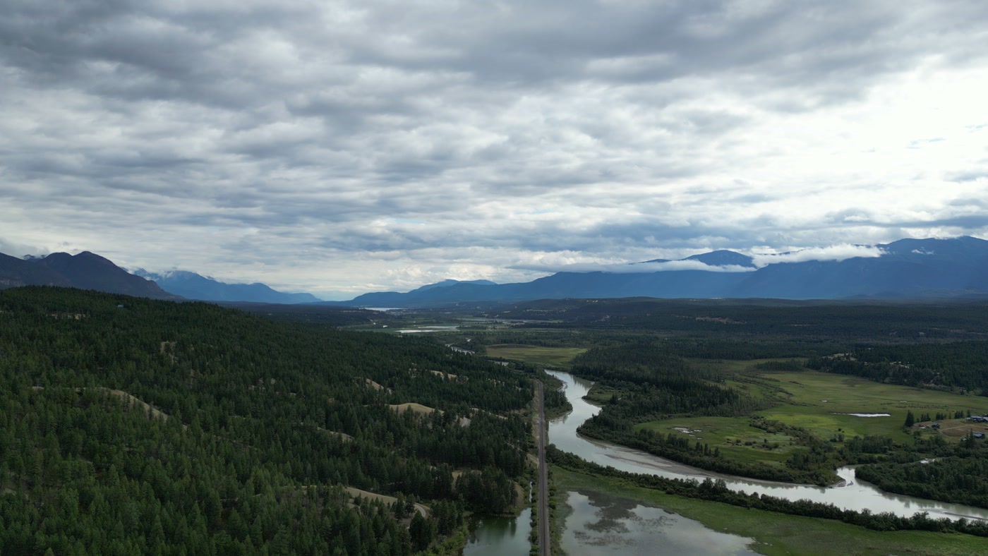 Down the Columbia Valley from above a forested slope near Radium Hot Springs: the river with the rail line on its bank running toward the far ranges, fields to the right, a dark peak close on the left