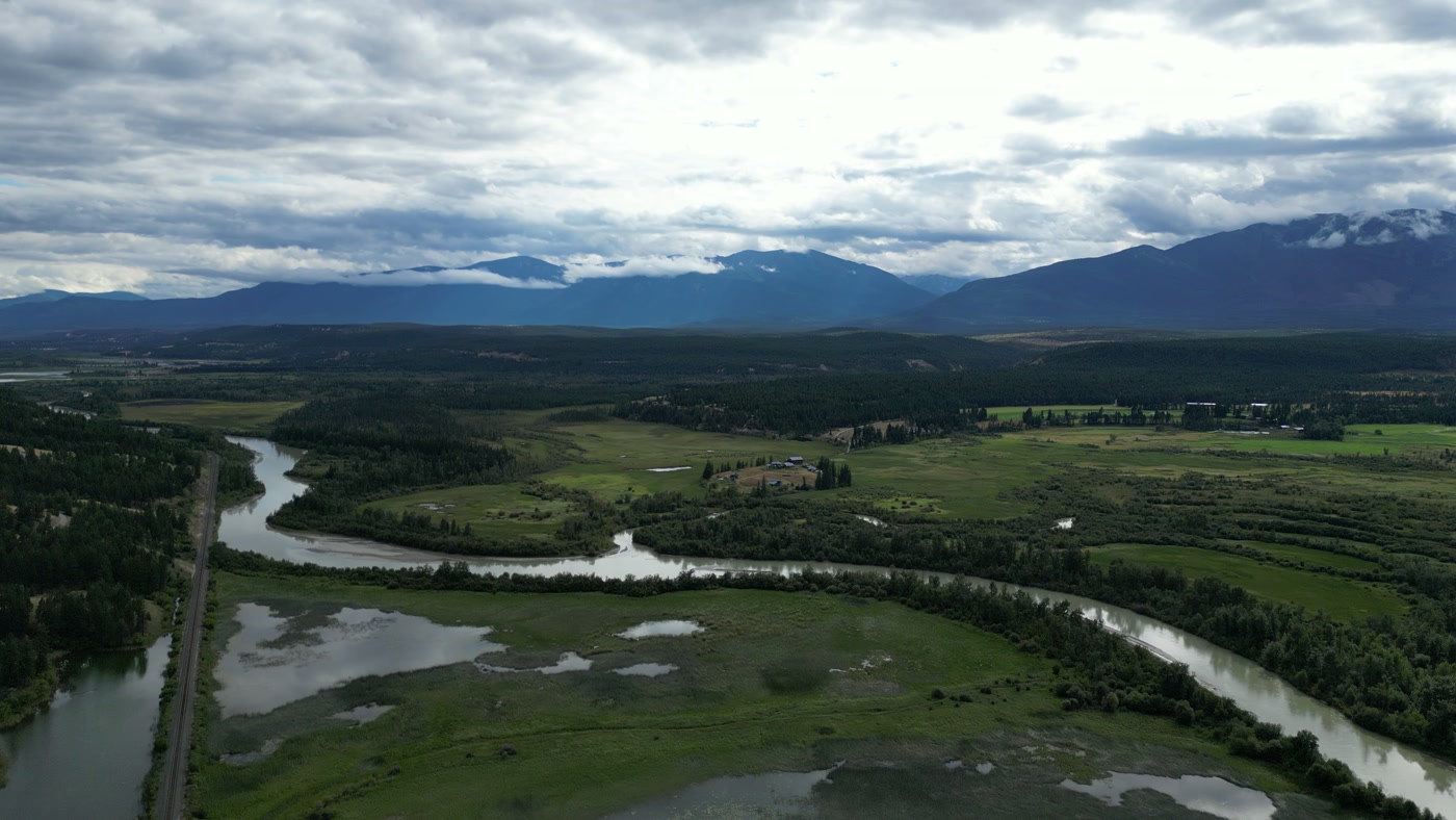 The Columbia River's S-bend through the wetland at Radium Hot Springs, the railway beside it, oxbow ponds, a farmstead on the bench and both ranges under cloud