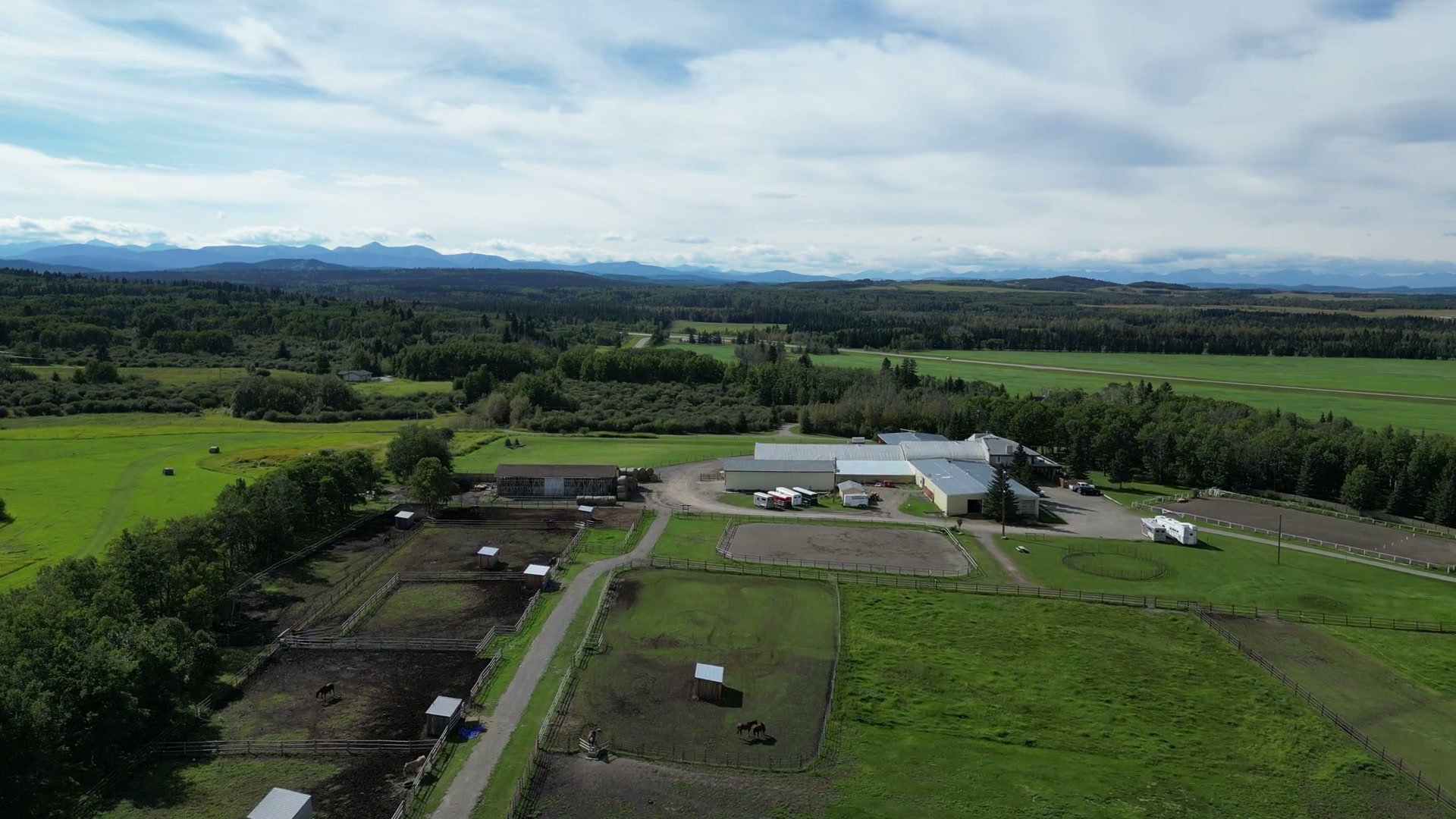 High over the paddocks of Shire Ridge Ranch near Bragg Creek: shelters dotted across the green, sand rings, the white arena buildings and trailers, and the mountains along the horizon