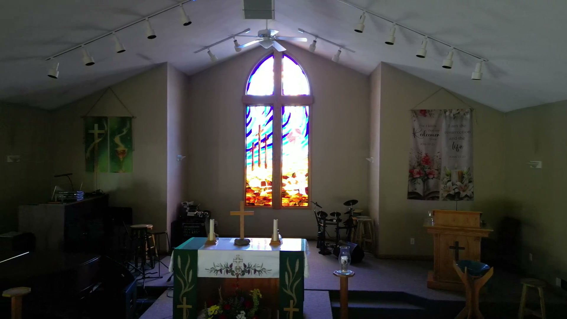 The chancel at St. Peter's held in frame: the window in the gable, a green banner to one side and a flowered banner to the other, the table, the pulpit and the drum kit