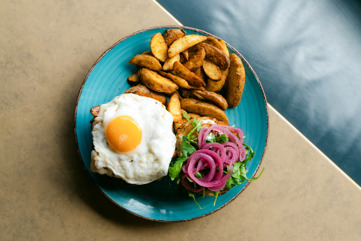 Sunny's Burger at Sunny Side Up in Cochrane on a turquoise plate, served open: a sunny-side-up egg over the patty, pickled red onions and arugula on the other half, herb potato wedges