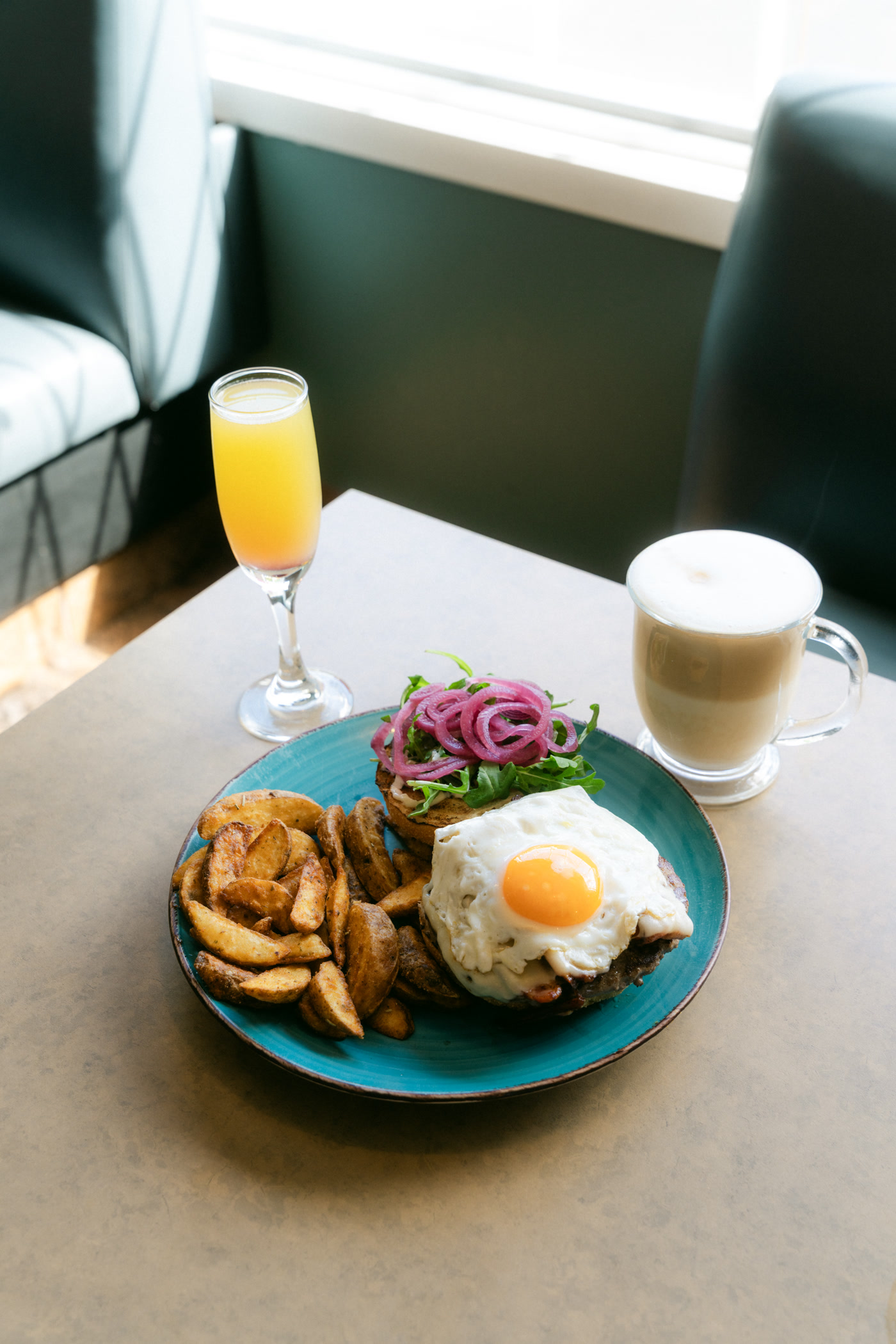 An orange drink in a tall stemmed flute and a glass mug of something milky either side of Sunny's Burger at Sunny Side Up in Cochrane, a teal booth behind