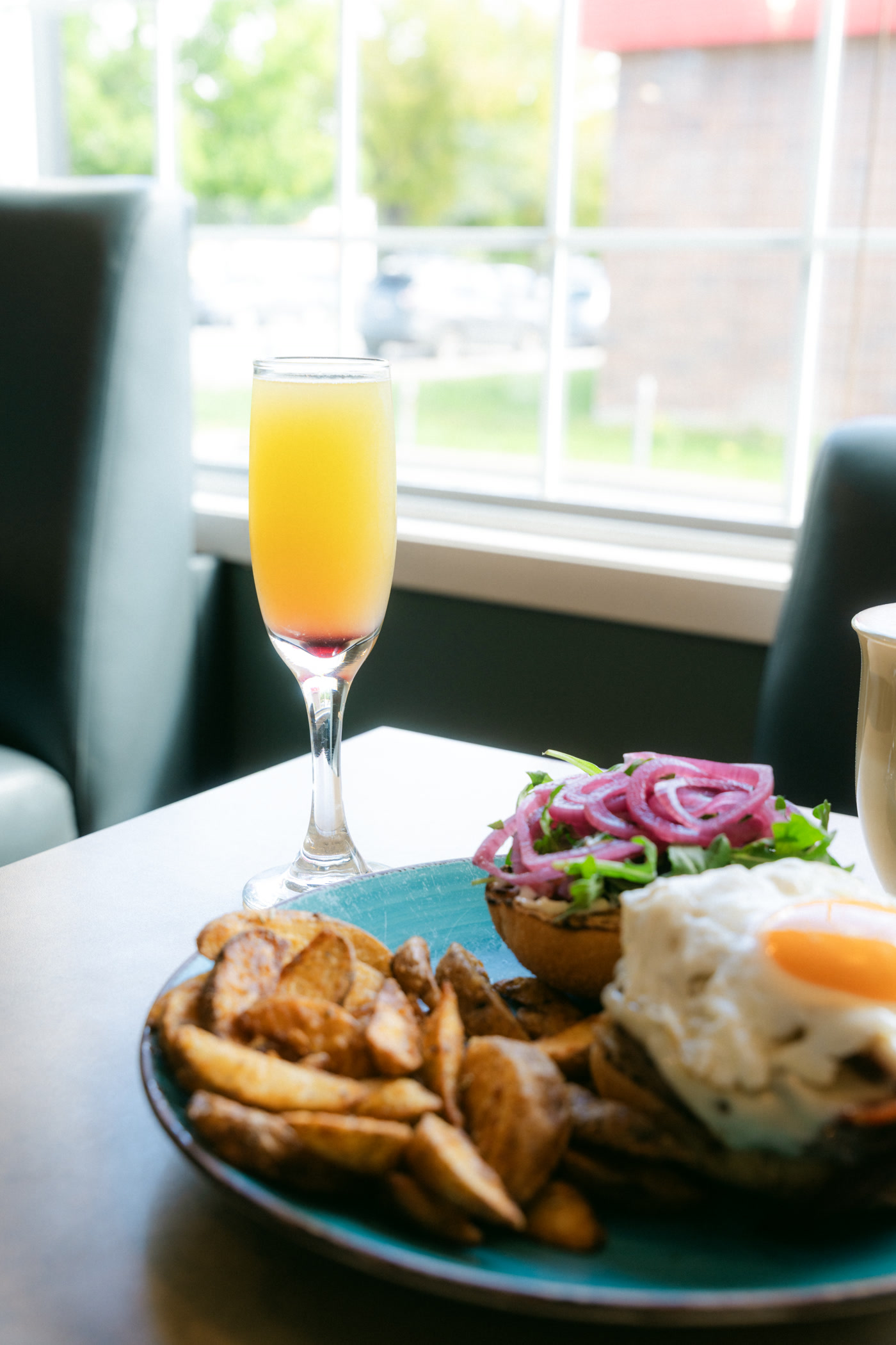 A flute of orange drink blushing red at its base, the burger and its wedges in the foreground, the window at Sunny Side Up in Cochrane and a red-trimmed brick building outside