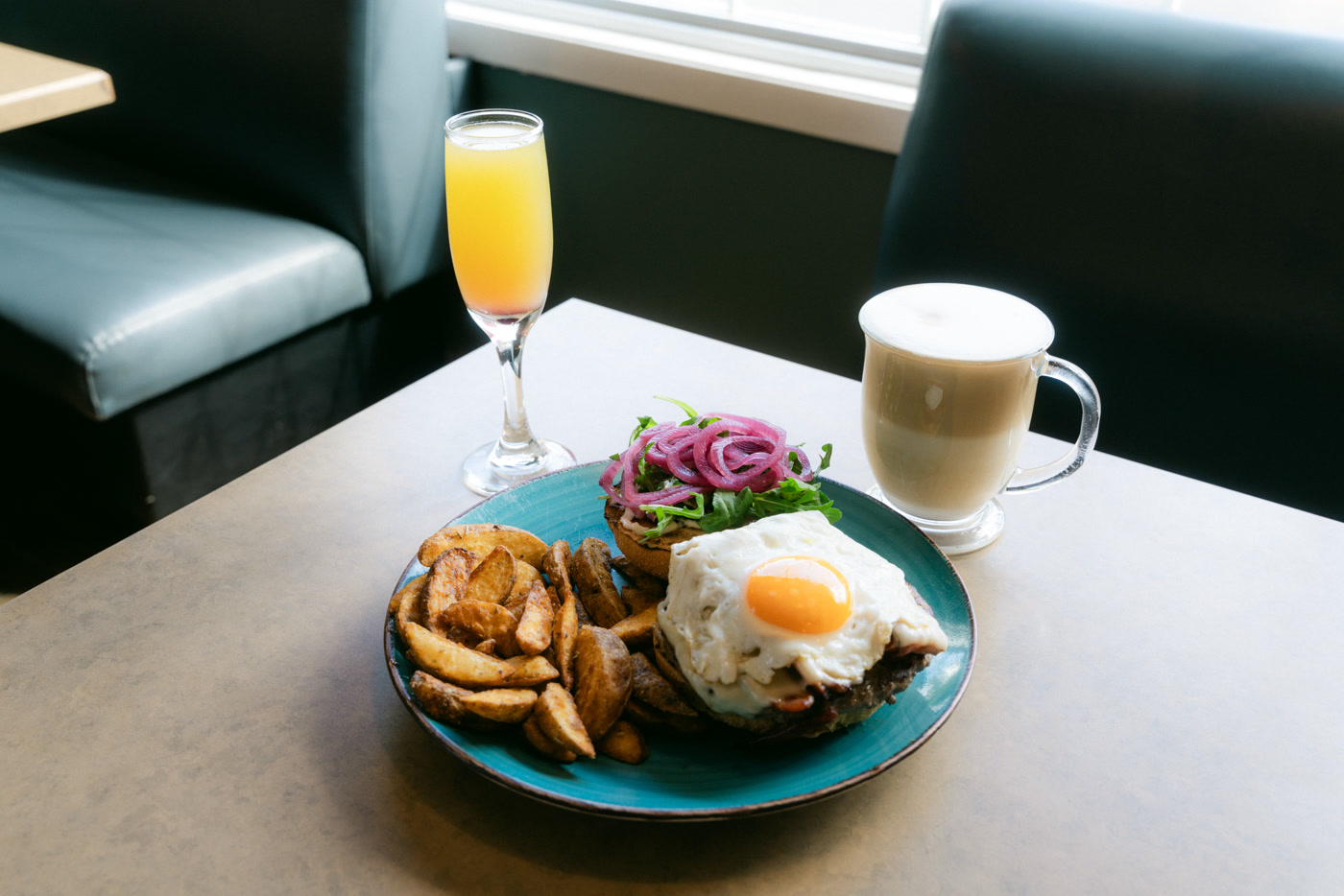 The whole table at Sunny Side Up in Cochrane between two teal booths: the burger on its turquoise plate, an orange flute, a glass mug and the window behind