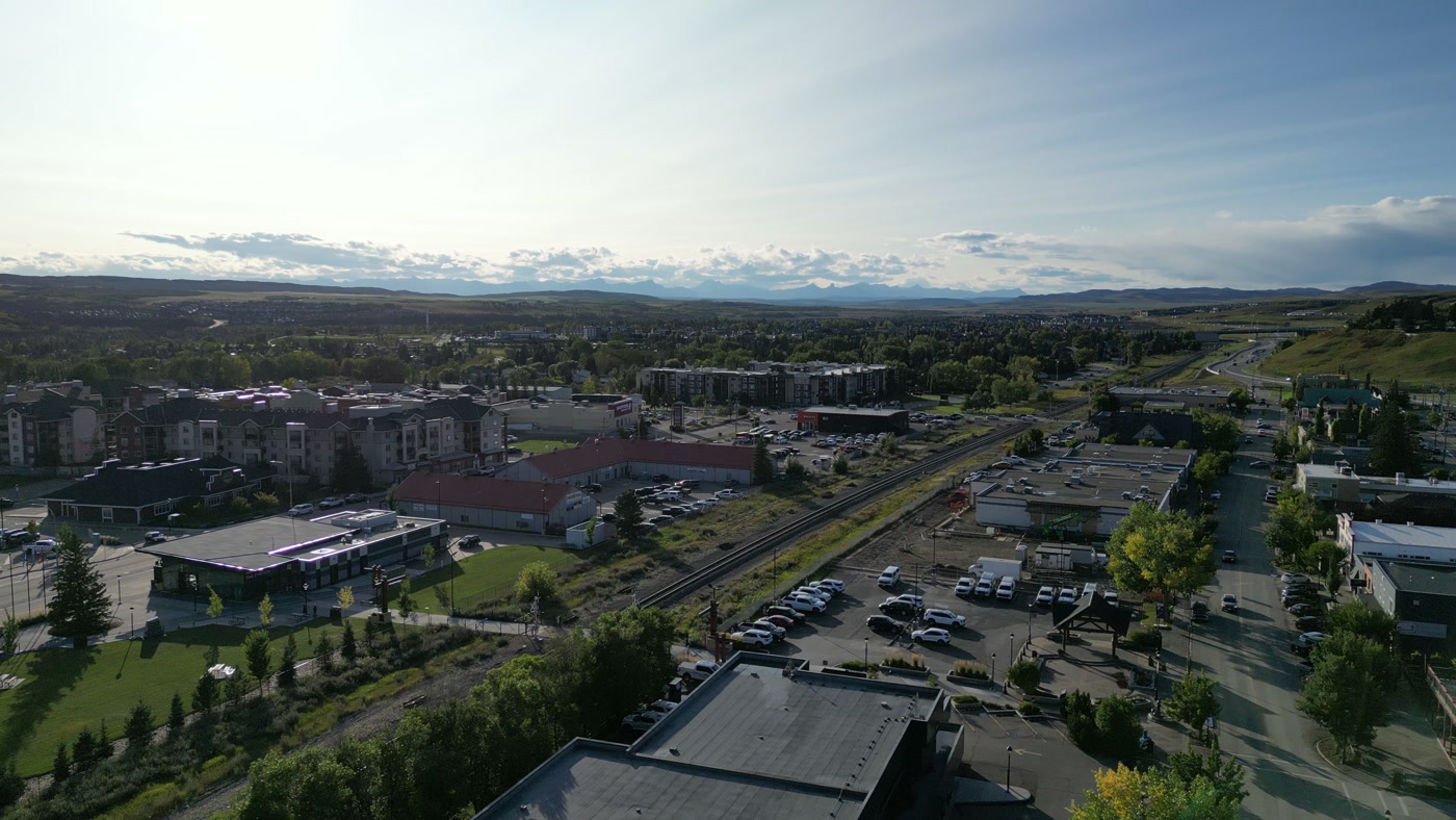 Past the rooftops of 1st Street West in Cochrane: the tracks curving away, parked cars, apartment blocks, low commercial buildings, green lawns and a long line of hills under a pale sky