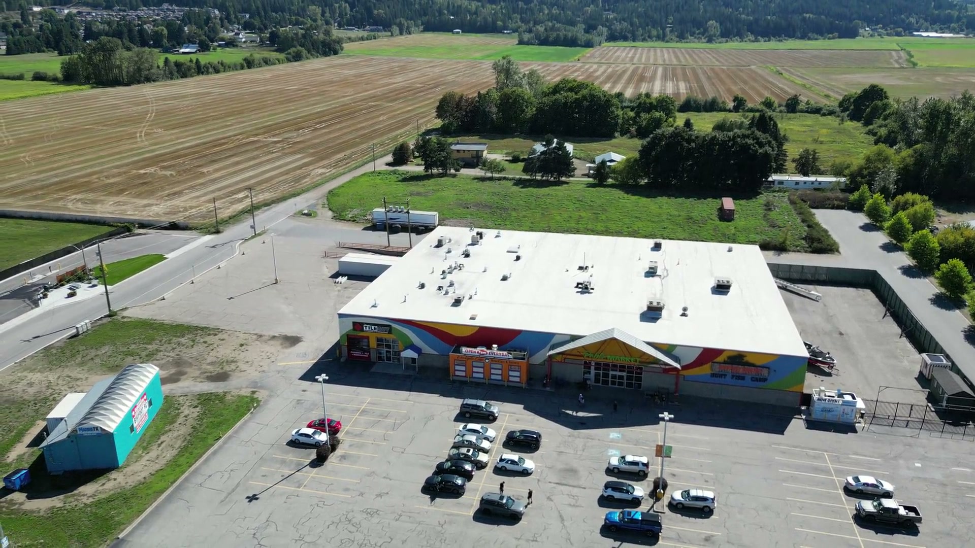 A slow orbit high above Westgate Public Market in Salmon Arm: the flat white roof, the painted front, rows of parked cars, a teal hut, and fields running back to a line of trees