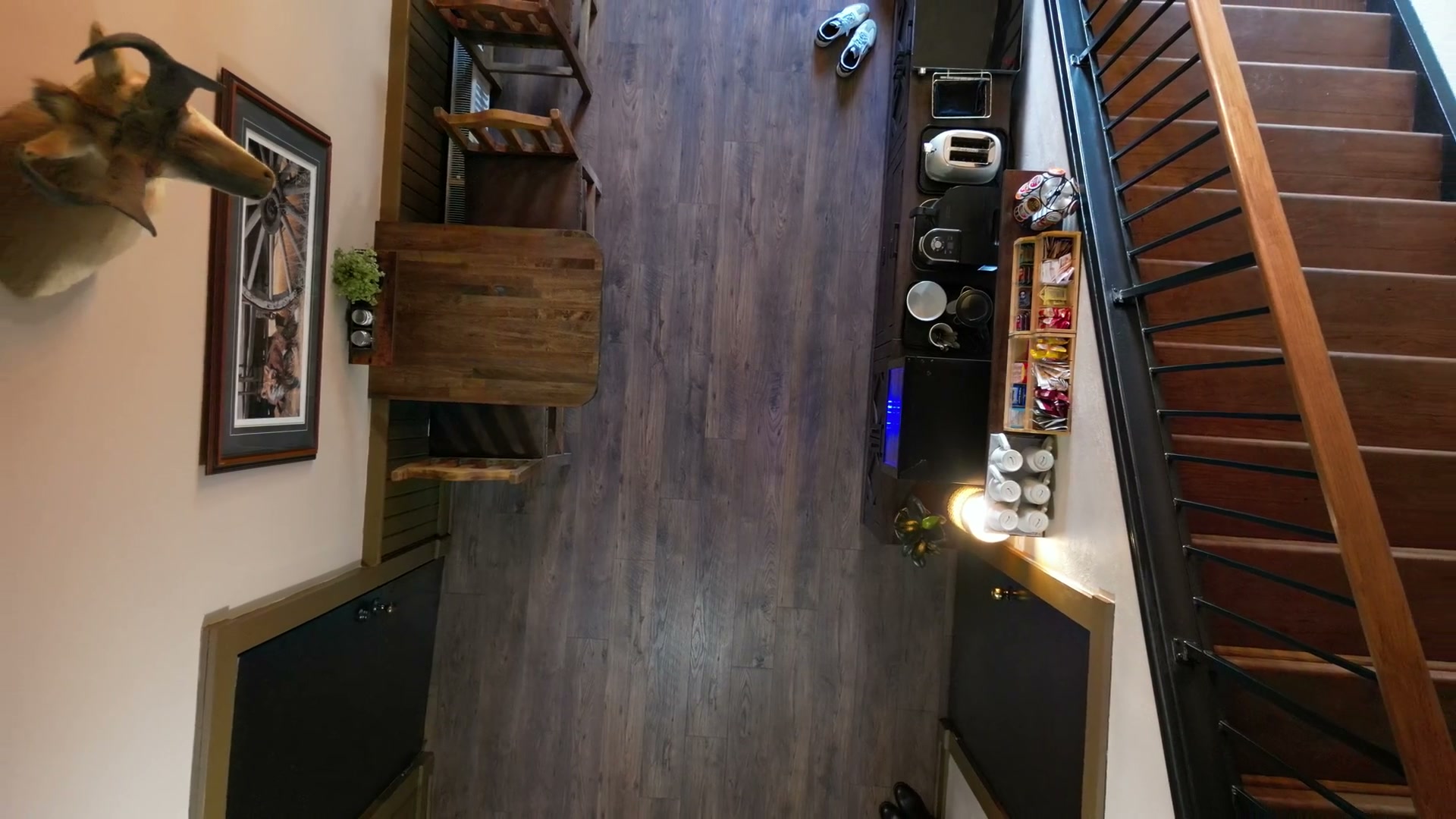 Looking straight down inside Shire Ridge Lodge near Bragg Creek over a wooden table and the coffee station, drifting until the staircase and a mounted head with curled horns come into the frame
