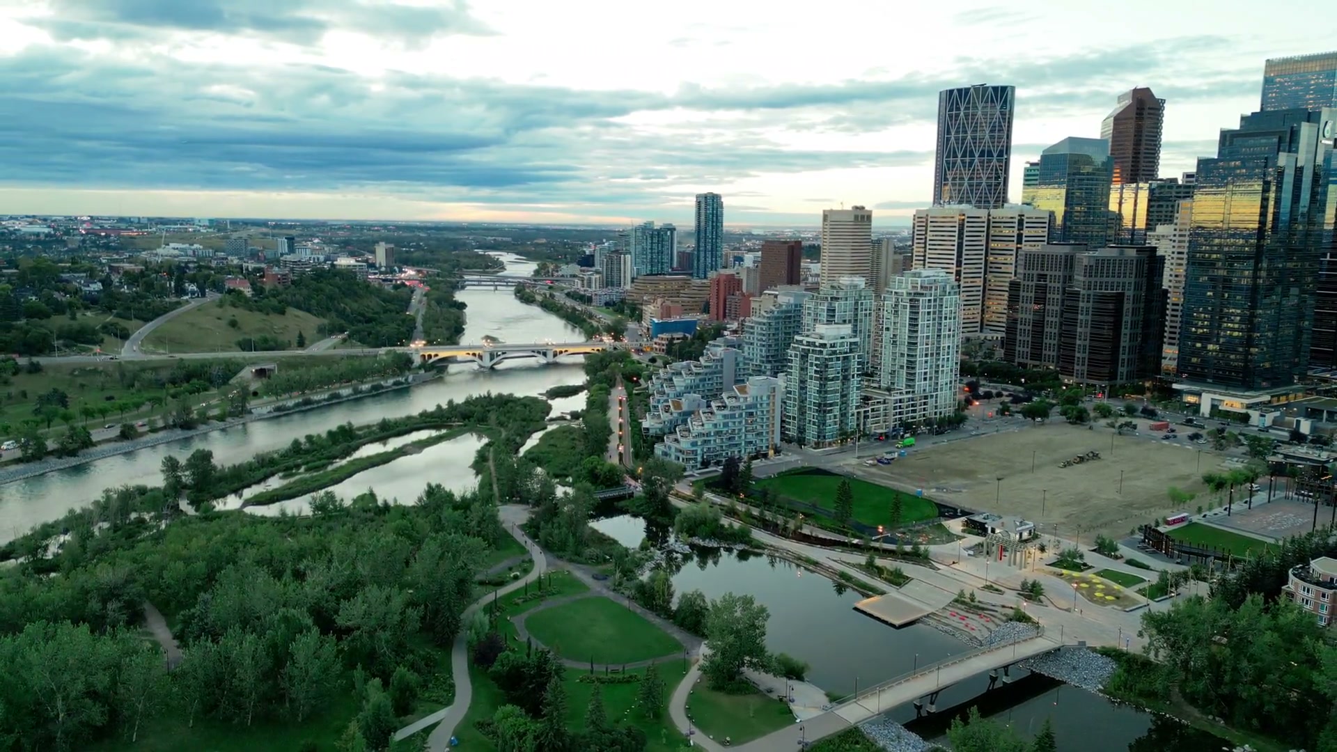 Descending toward the east end of Prince's Island at dusk: the wetland ponds, the footbridge and lit promenade to Eau Claire, downtown's towers above