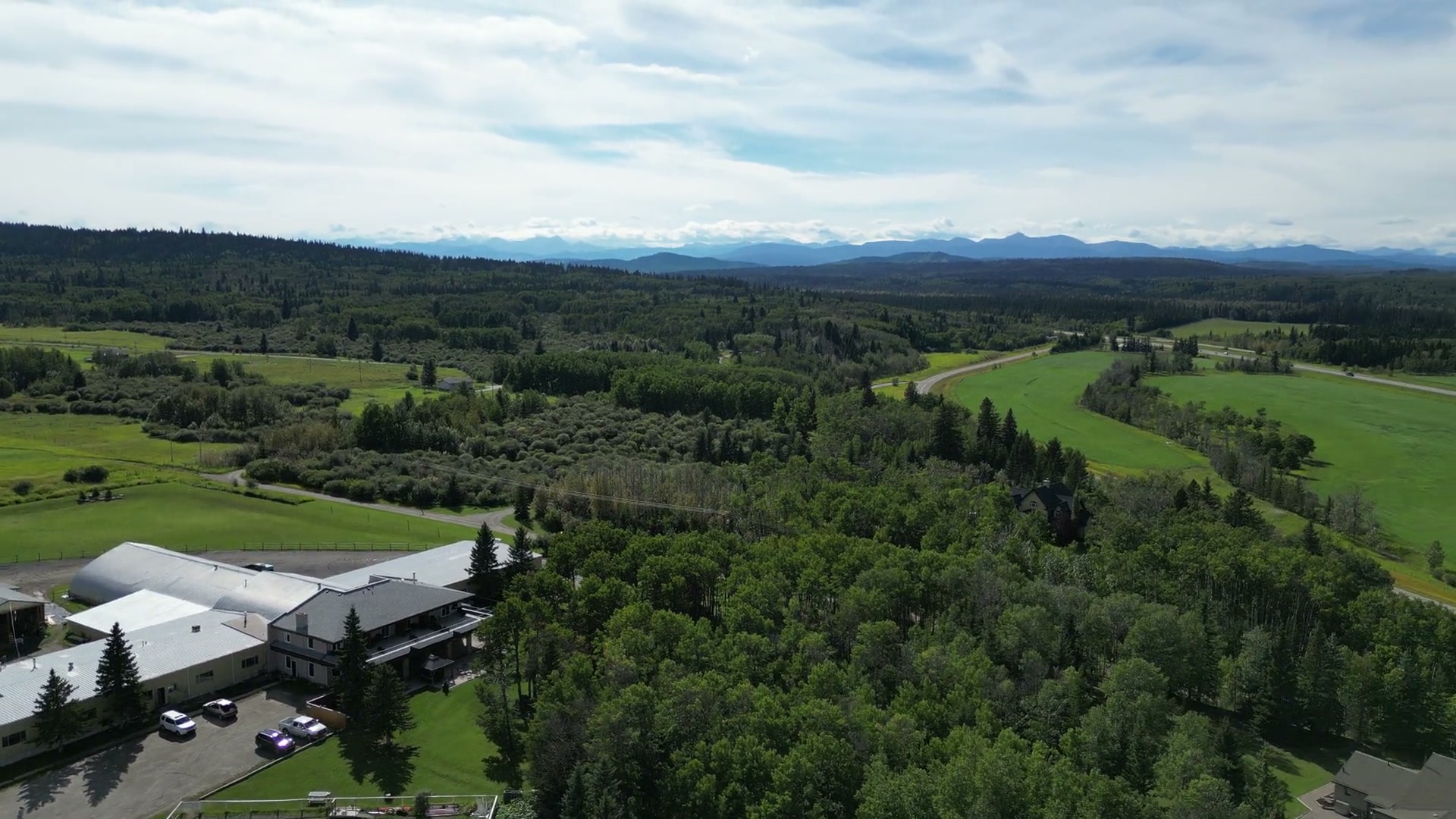 Rising from the lodge's wide deck at Shire Ridge Ranch near Bragg Creek over the white barn buildings, green fields and forest until the mountains line the horizon under the cloud