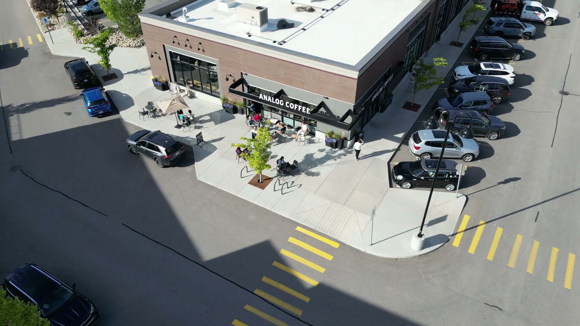 The cafe's corner unit from above: the patio, the yellow crosswalk bars, the parked cars, then the highway along the top of the frame