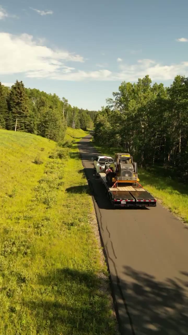 Aerial following a flatbed trailer carrying a compact track loader along a paved road through summer forest