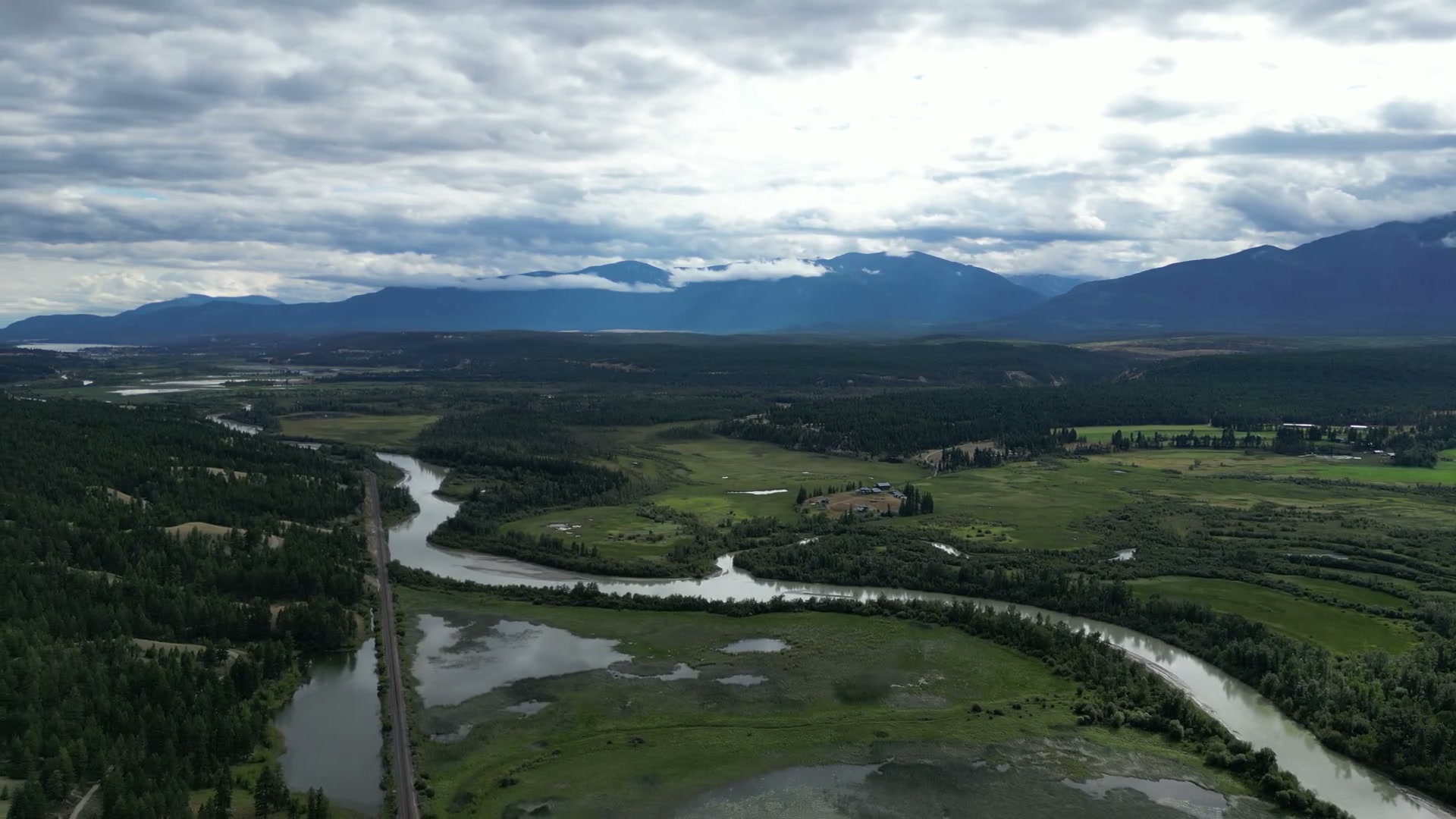 The top of the climb over the Columbia Wetlands: the river's bend, the railway, the fields and farm on the bench, and the ranges on both sides of the valley under a sheet of cloud