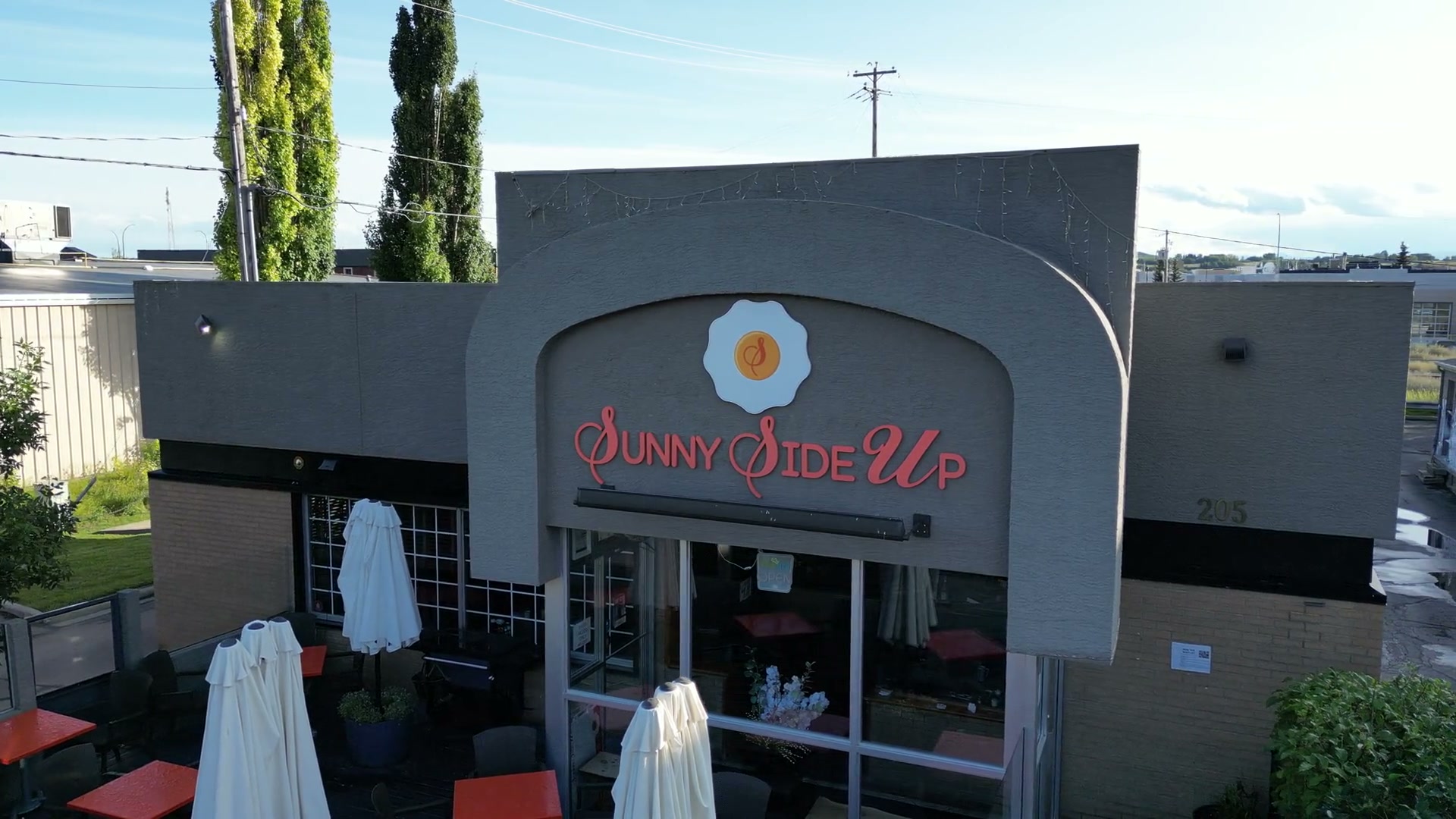 Sliding right along the front of Sunny Side Up in Cochrane, the patio's furled white umbrellas and red tables in the foreground, the arched sign and glass doors ahead, poplars behind