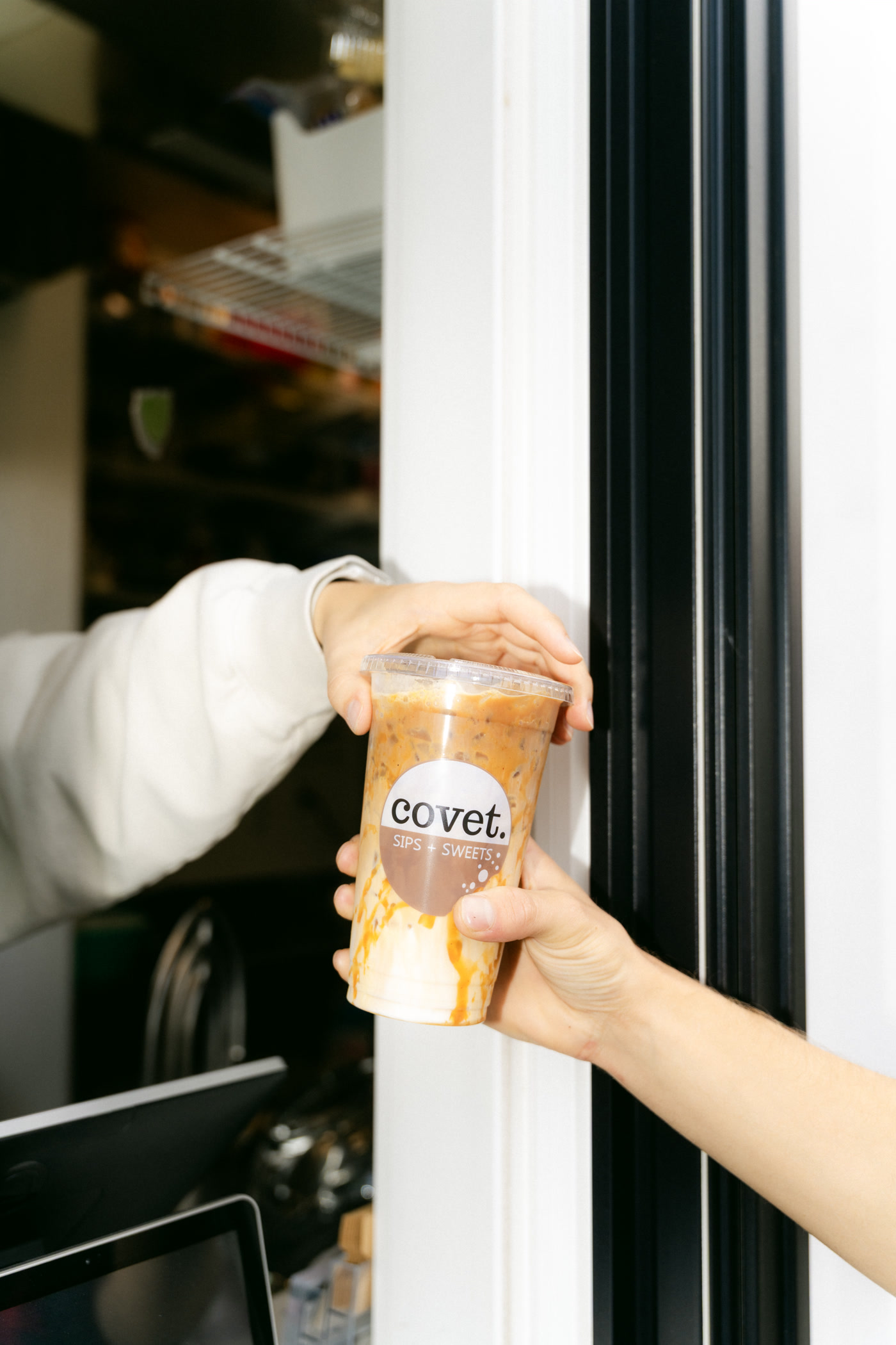 A caramel iced drink handed through the serving window of the Covet Sips + Sweets container in Cochrane, the till screen below it