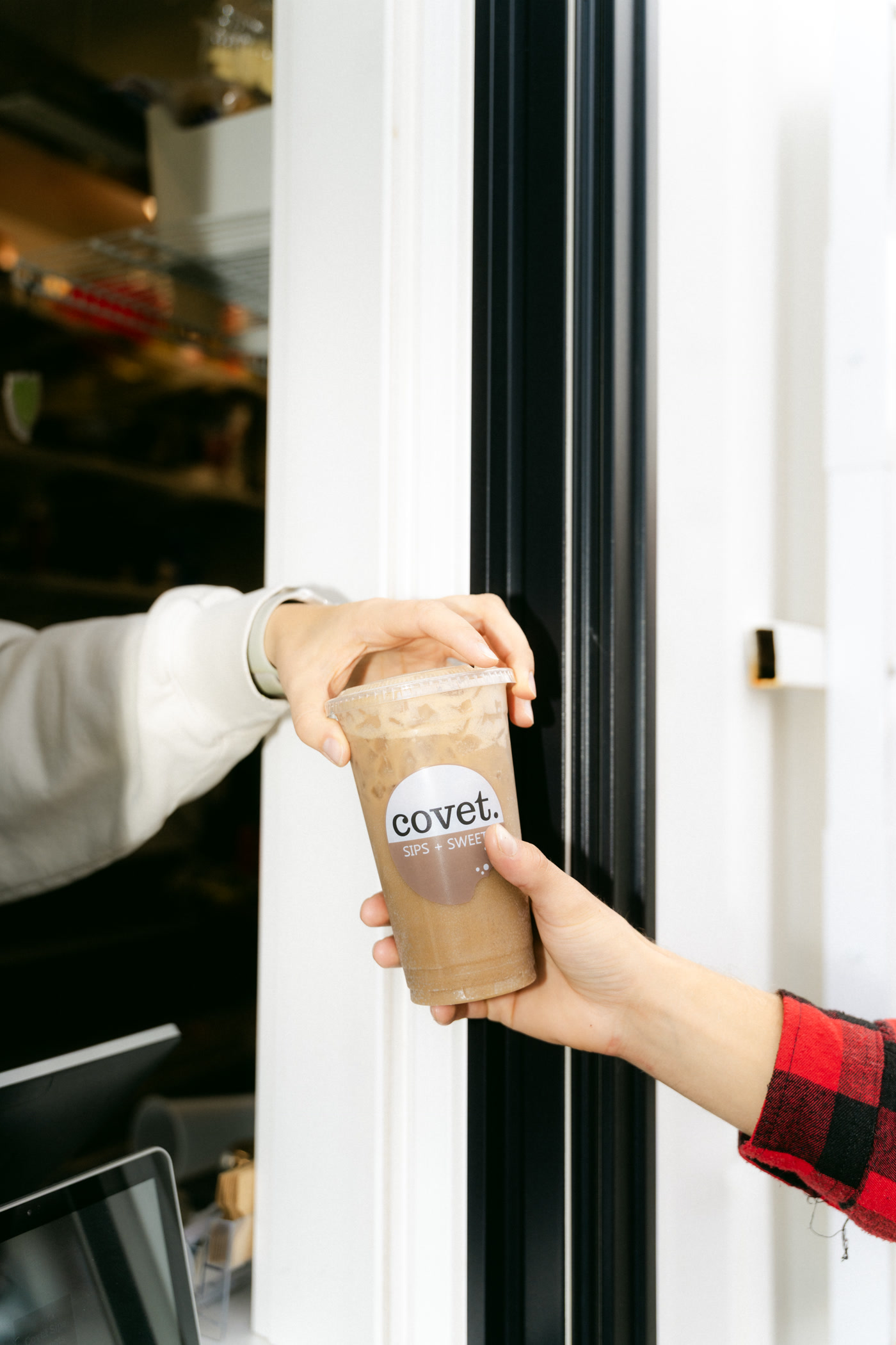 An iced coffee in a covet cup handed out through the serving window of Covet Sips + Sweets in Cochrane to a hand in a red plaid sleeve
