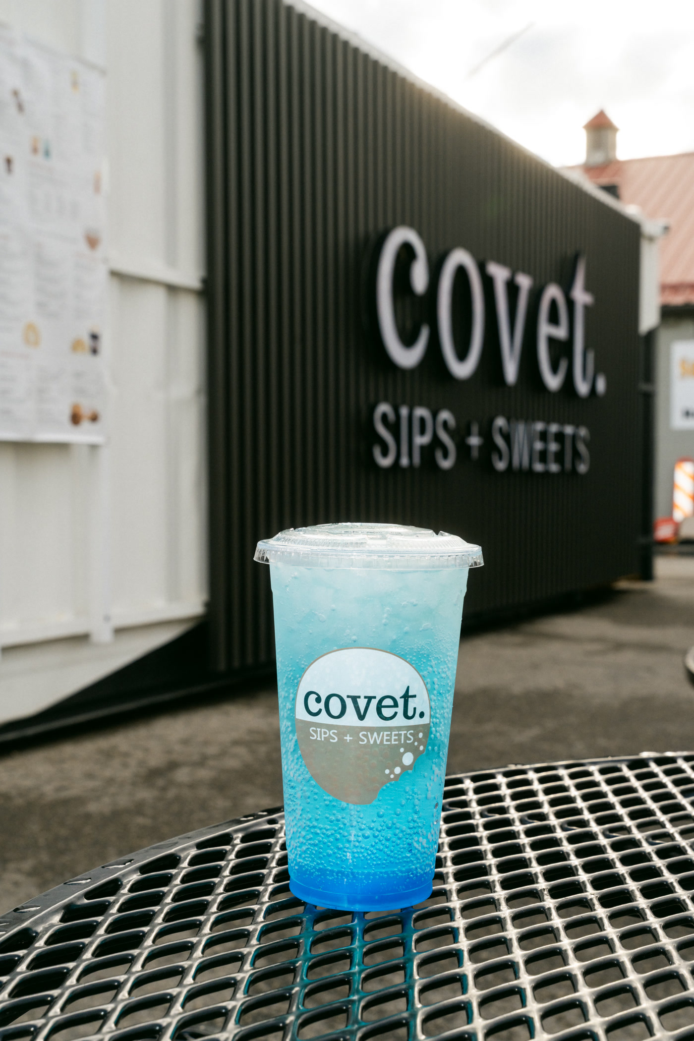 A tall blue layered drink in a covet cup on the black mesh patio table outside Covet Sips + Sweets in Cochrane