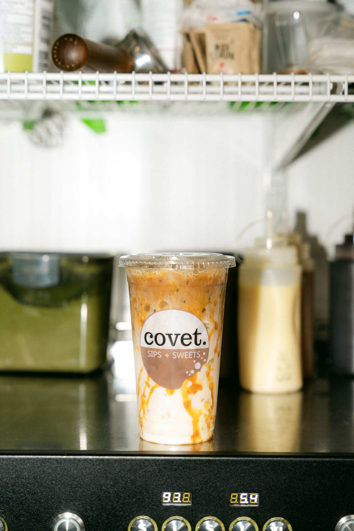 A caramel-drizzled iced drink in a covet cup on top of the espresso machine at Covet Sips + Sweets in Cochrane, a wire shelf above it