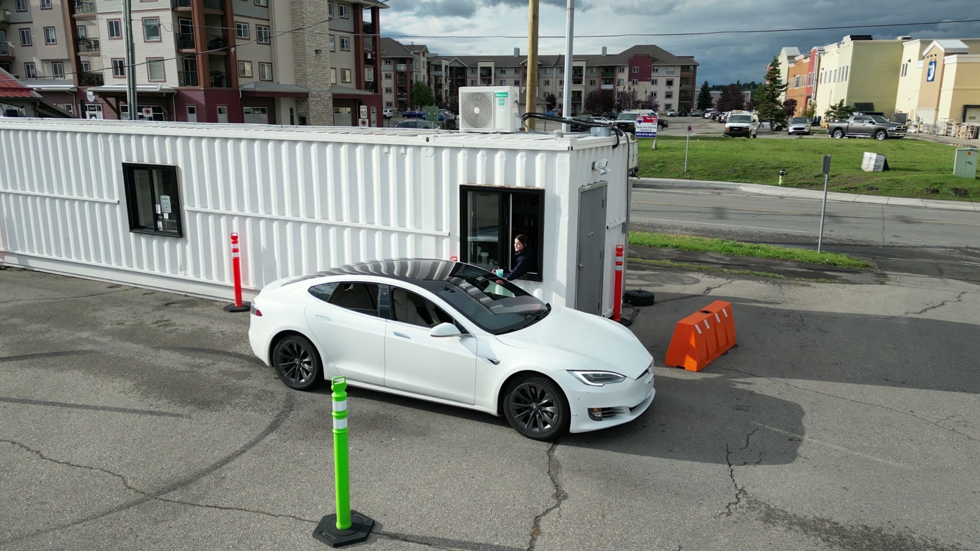 A white car at the end window of the Covet Sips + Sweets container in Cochrane, someone inside leaning out toward it, the road and a green lot beyond