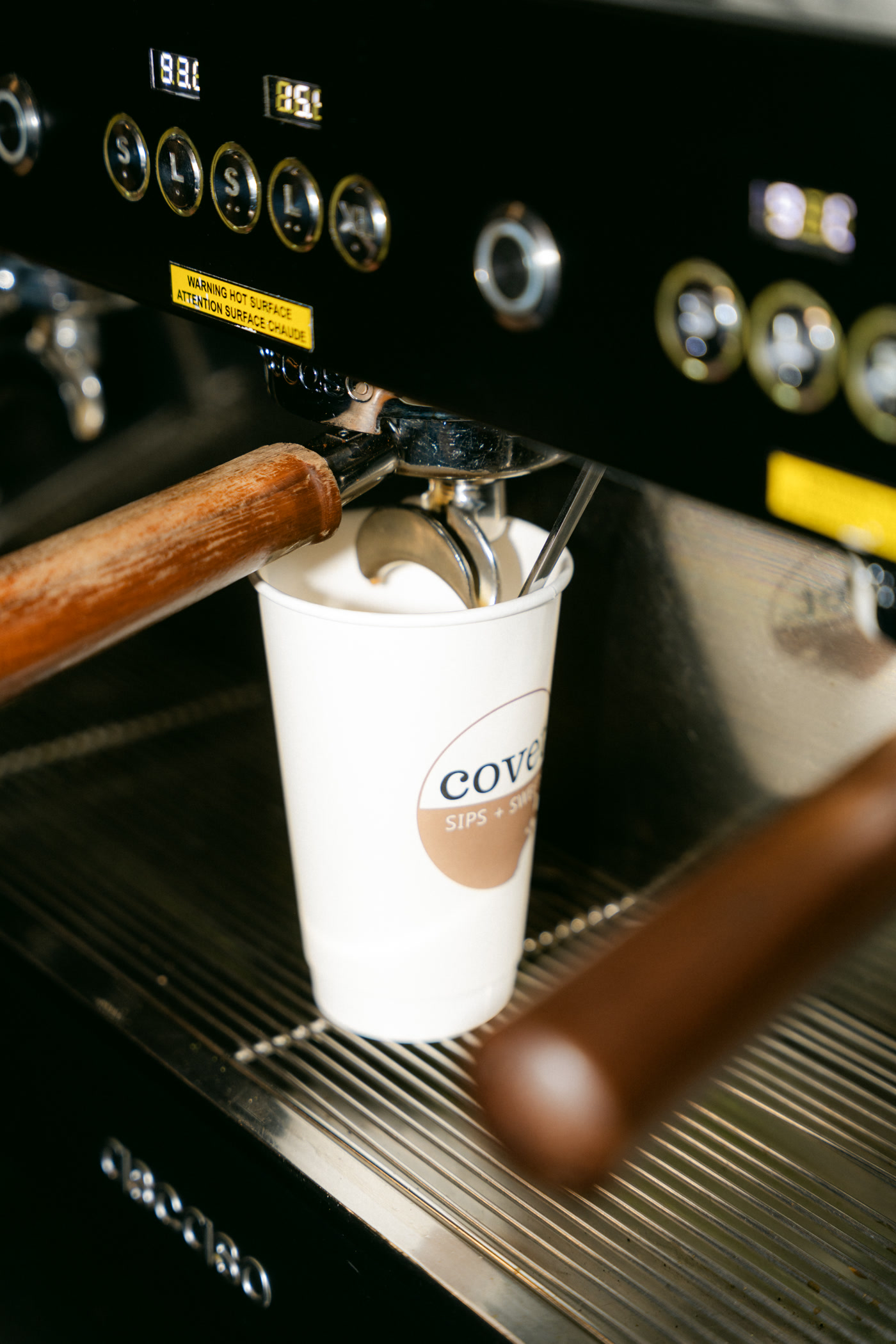A white covet paper cup standing under the espresso machine at Covet Sips + Sweets in Cochrane, a wooden-handled portafilter either side