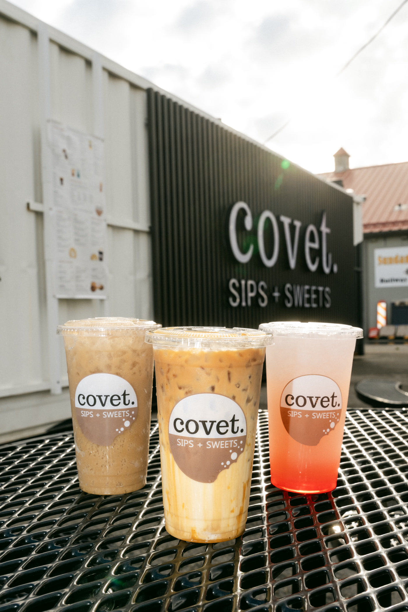 Three iced drinks in covet cups on a black mesh patio table outside the Covet Sips + Sweets container in Cochrane, the sign wall behind
