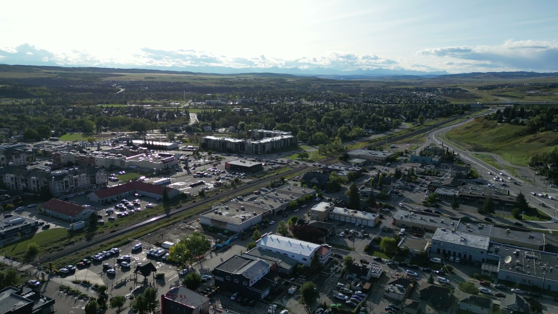 Rising over downtown Cochrane as the view widens: flat roofs and parking lots, apartment blocks, a rail line cutting across the town, a highway curving past a green hillside, and the mountains a pale line