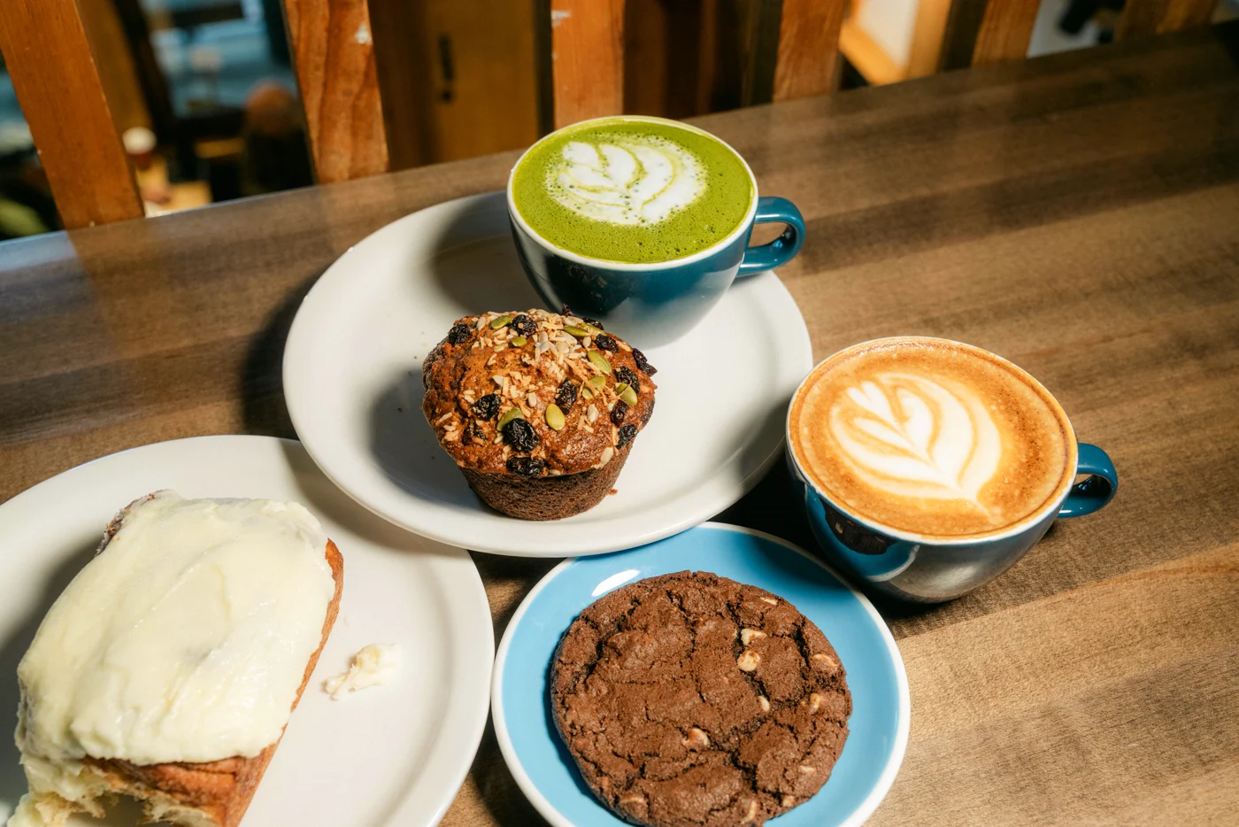 A café table from above: a matcha latte with leaf art, a muffin, a frosted bun, a chocolate cookie and a latte