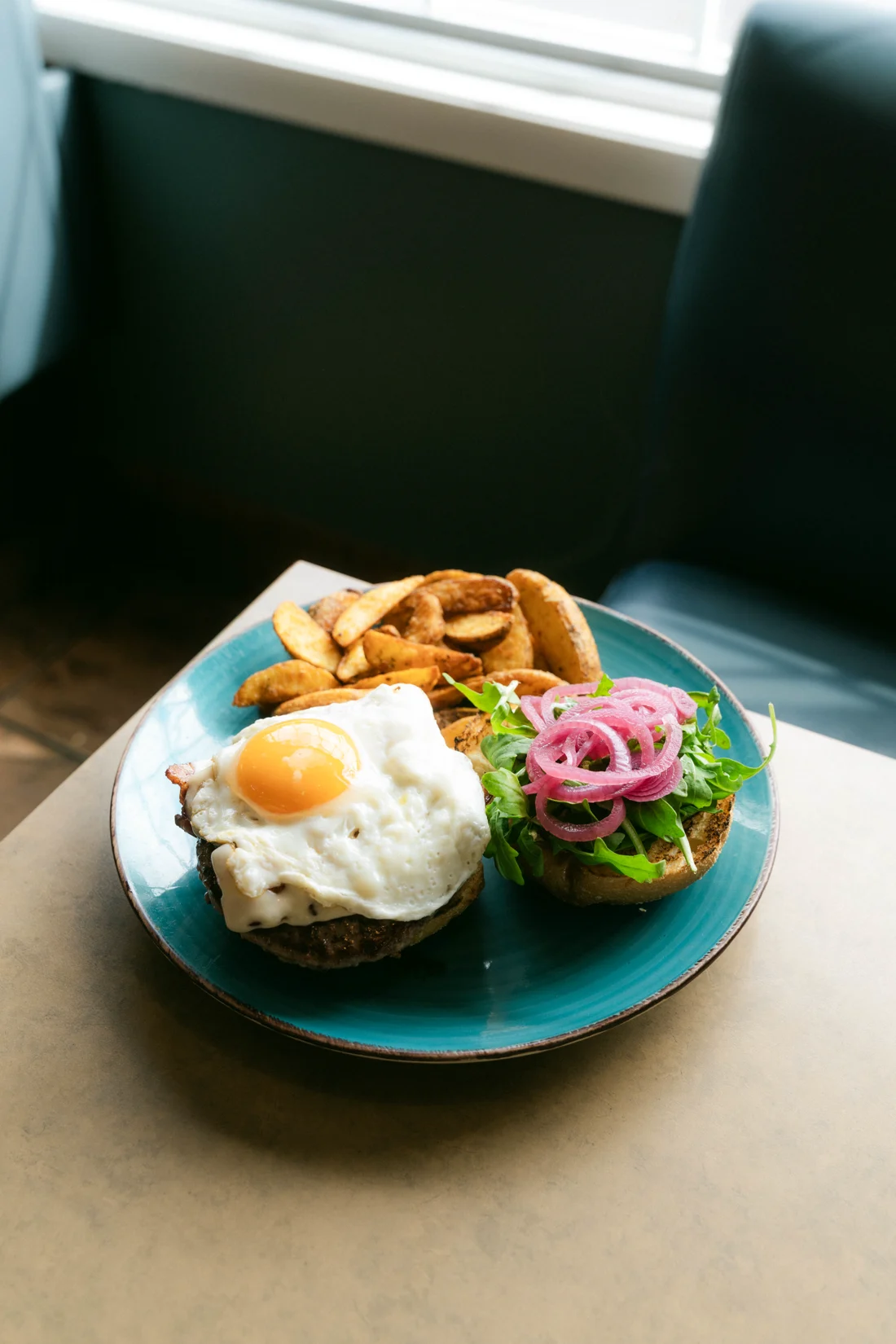 A burger topped with a fried egg and pickled onion, potato wedges beside it, on a teal plate in a diner booth