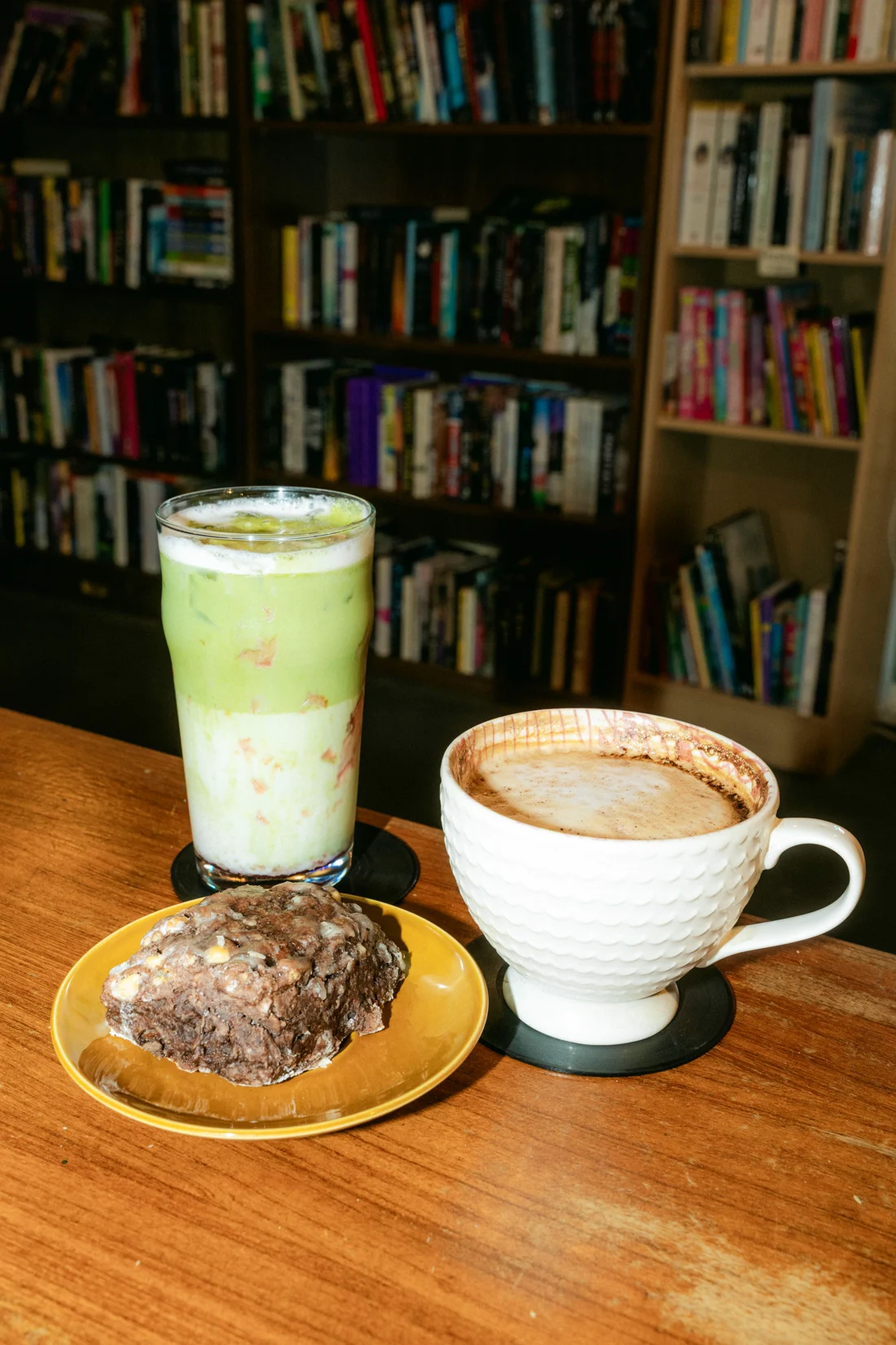 An iced matcha, a latte and a chocolate cookie on a wood table with bookshelves behind