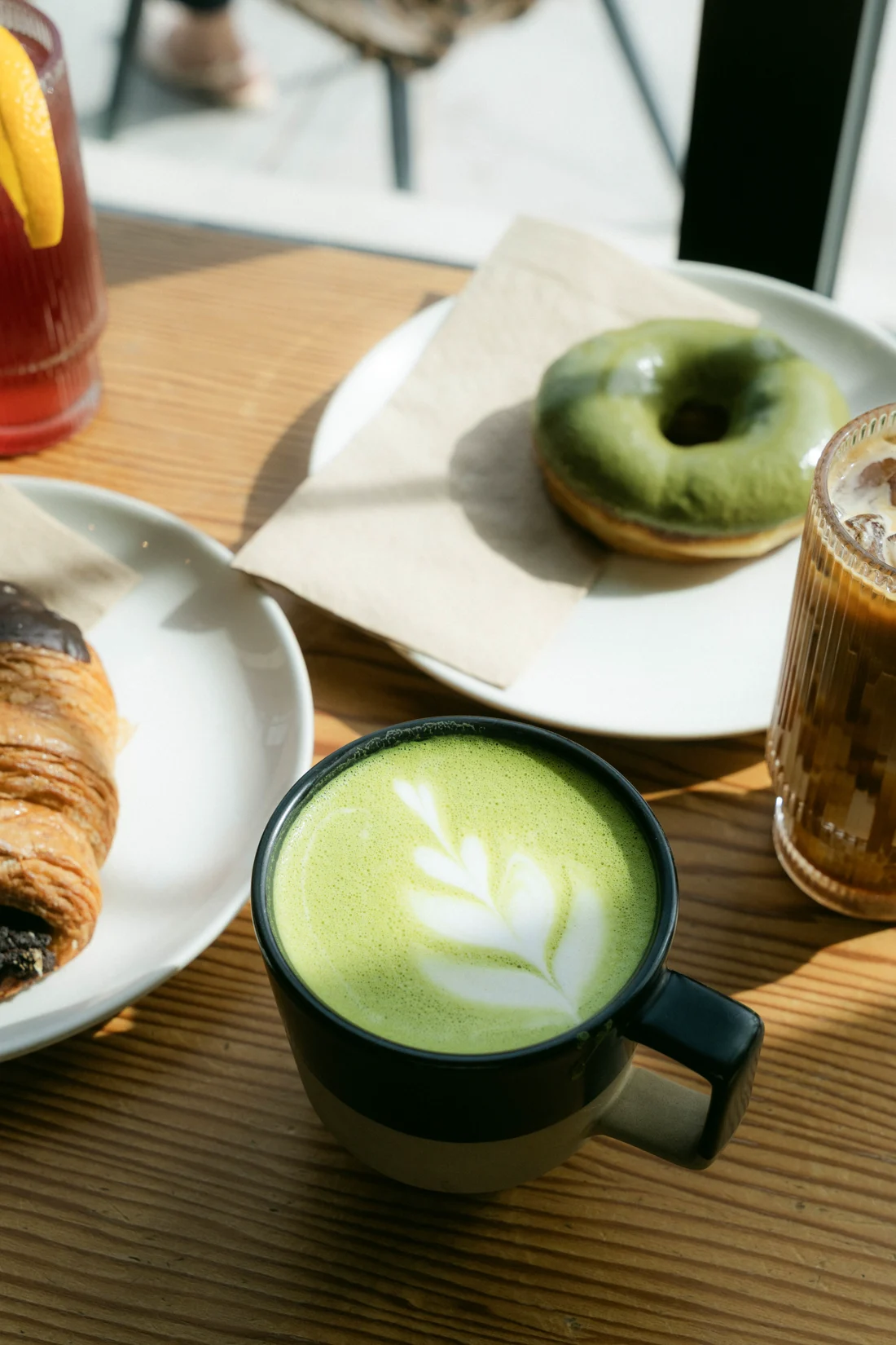 A matcha latte with a leaf poured in its foam beside a doughnut and an iced coffee