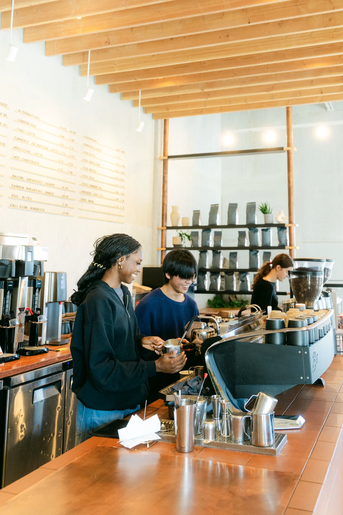 Two baristas work the espresso machine under a timber ceiling, a wall of jars behind them