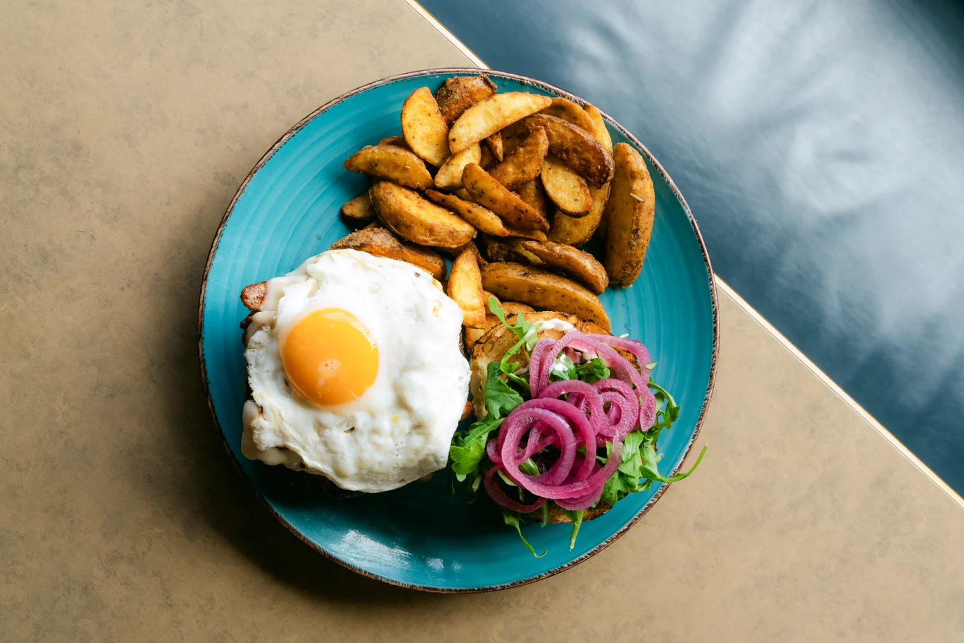 From above: a fried-egg burger, potato wedges and pickled onion on a teal plate