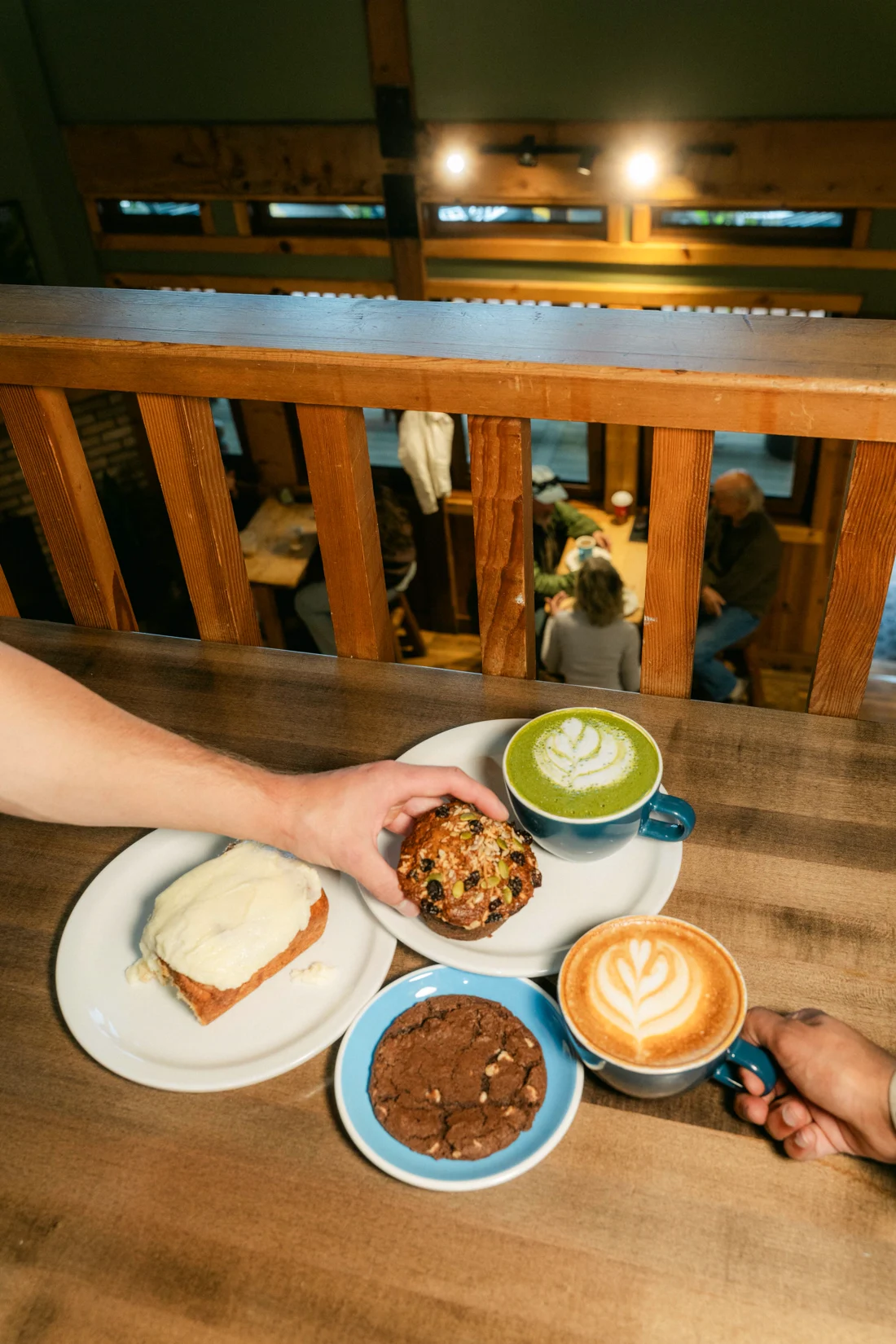 Hands reach across a wood table for a matcha latte, a muffin and a chocolate cookie, a café balcony behind