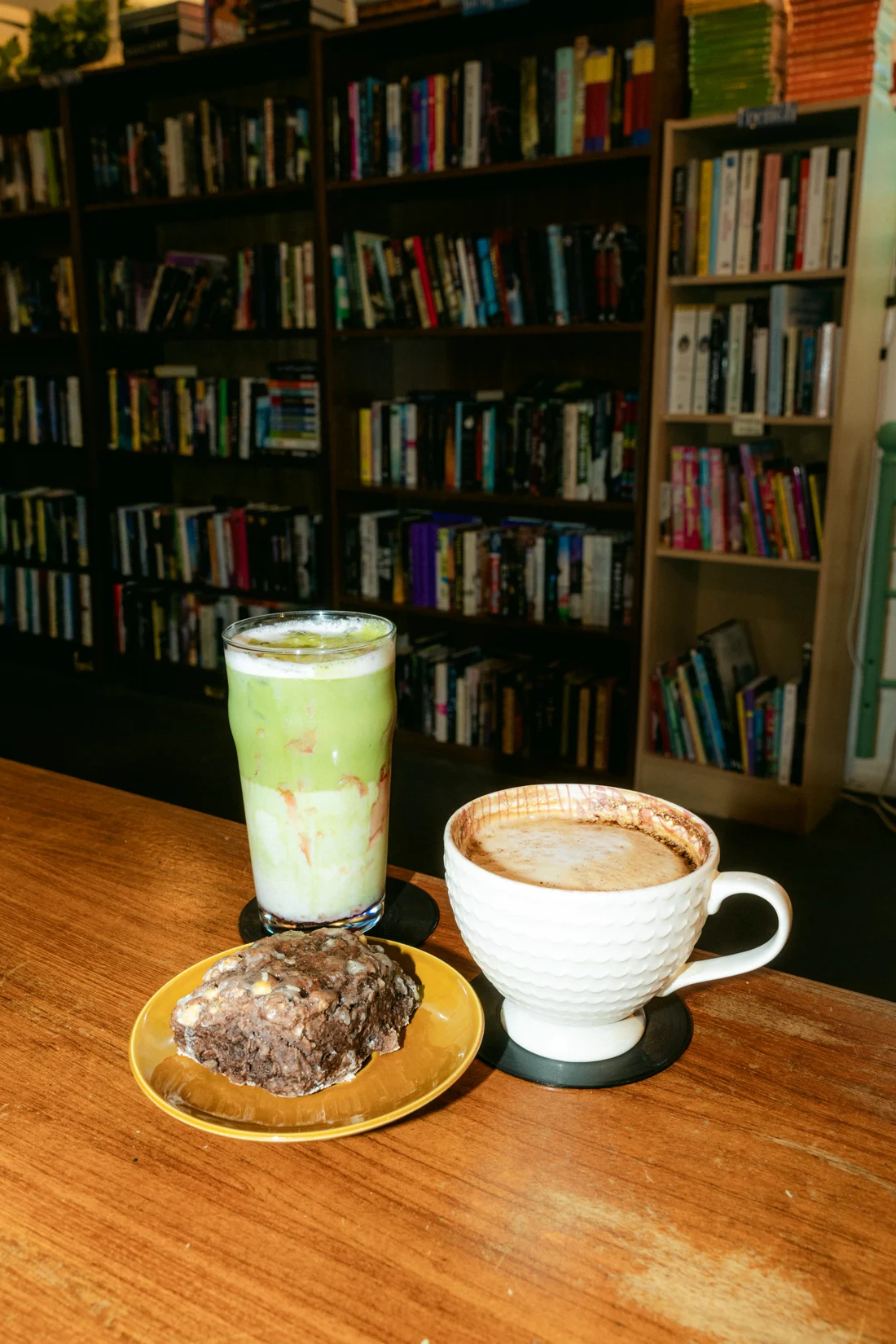 An iced matcha, a latte and a cookie on a table in front of bookshelves