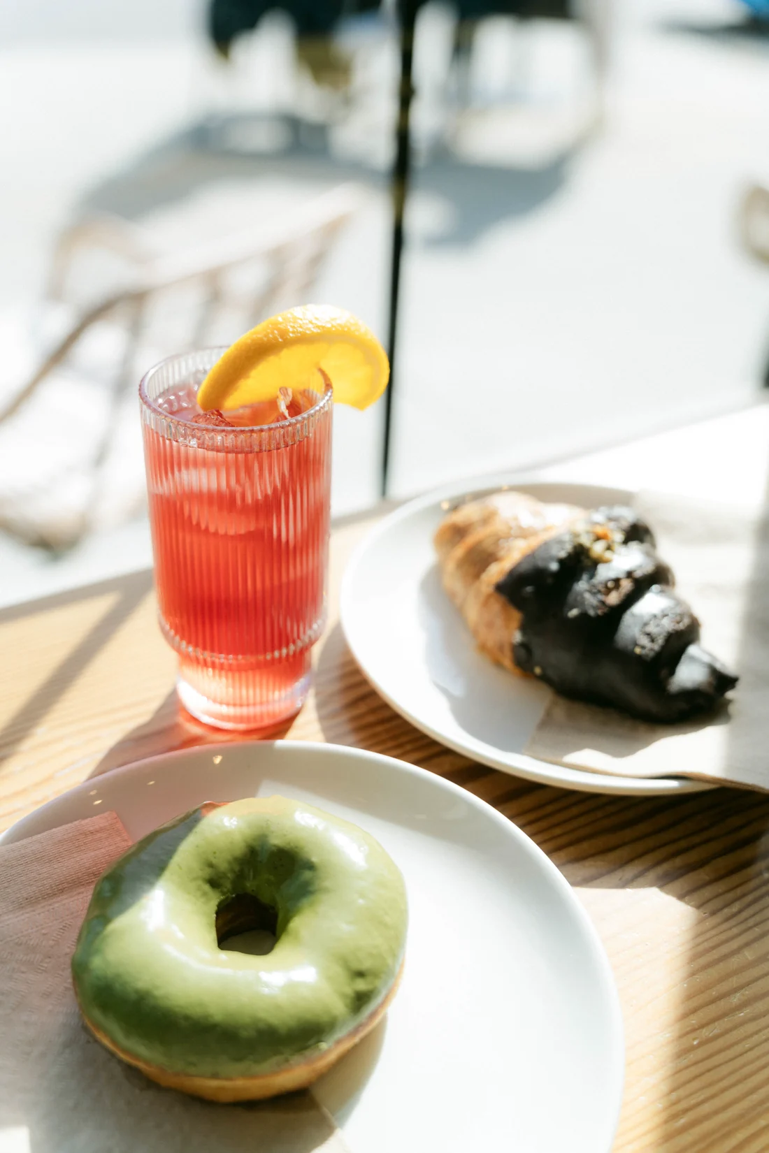 A red iced drink with an orange slice, a croissant and a matcha doughnut on a sunlit café table