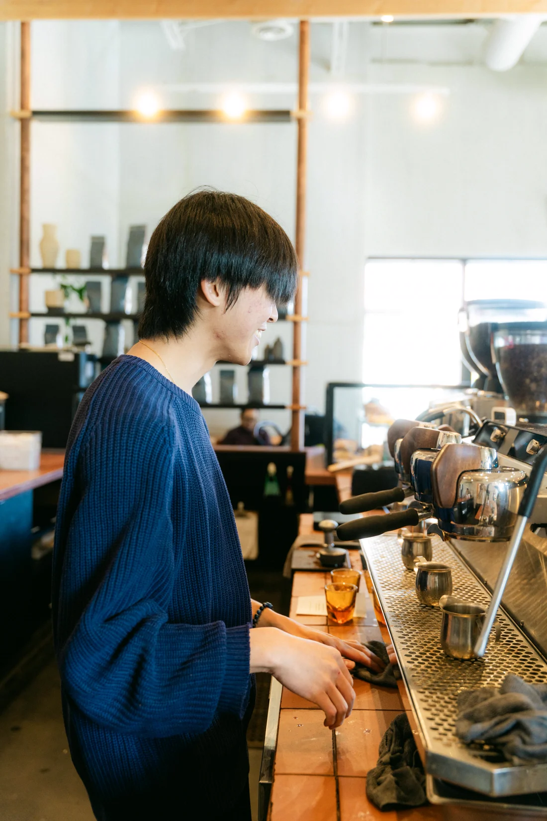 A barista in a navy sweater works the espresso machine in profile, jars on the shelves behind
