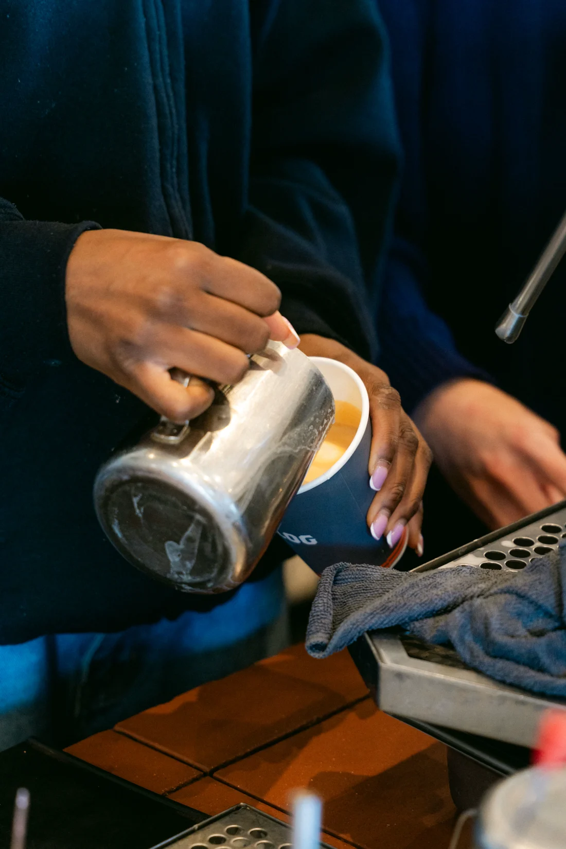 Hands pour steamed milk from a steel jug into a cup at the espresso machine