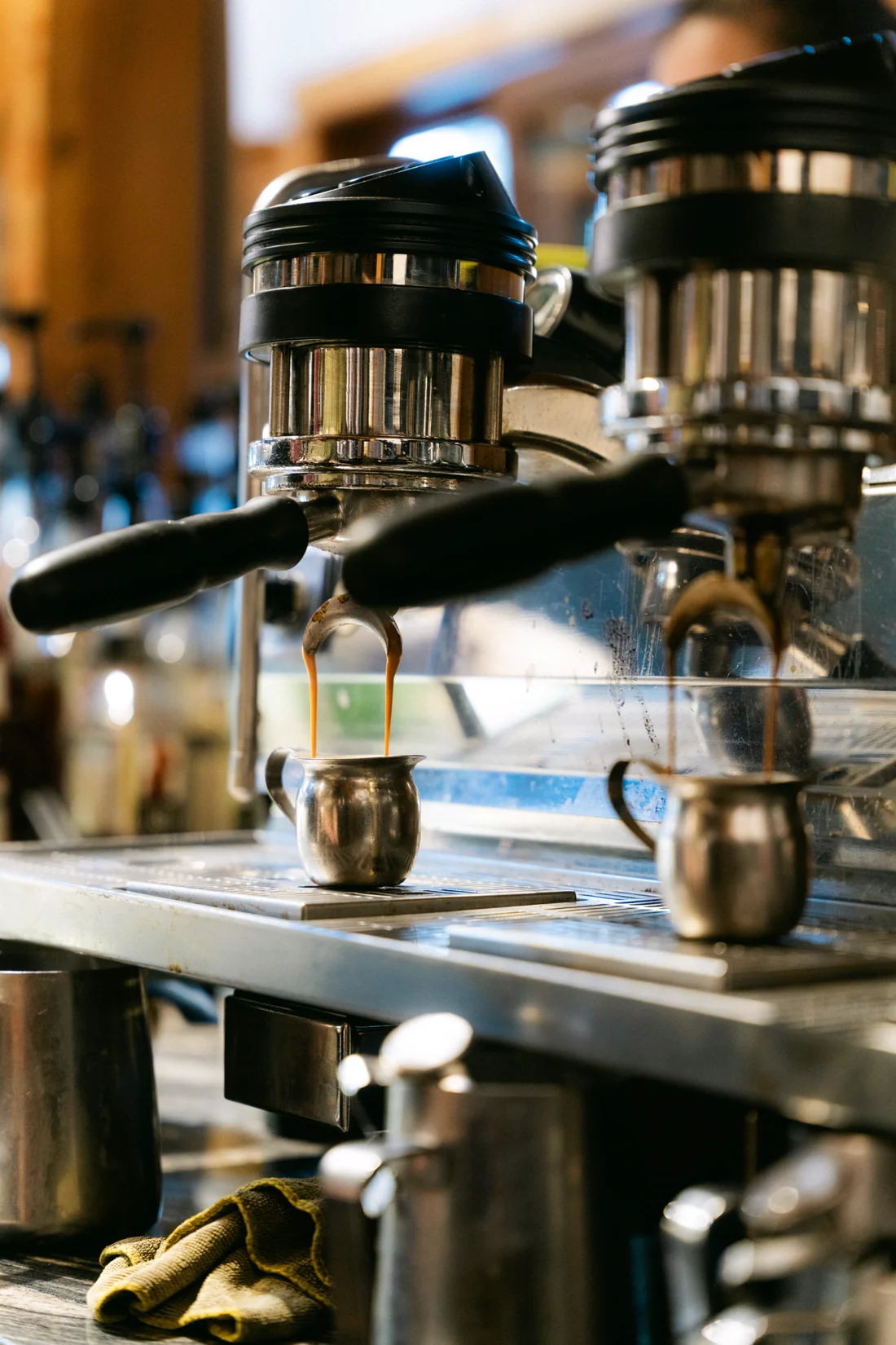 Two portafilters dripping espresso into small steel jugs on a chrome machine, the café blurred behind
