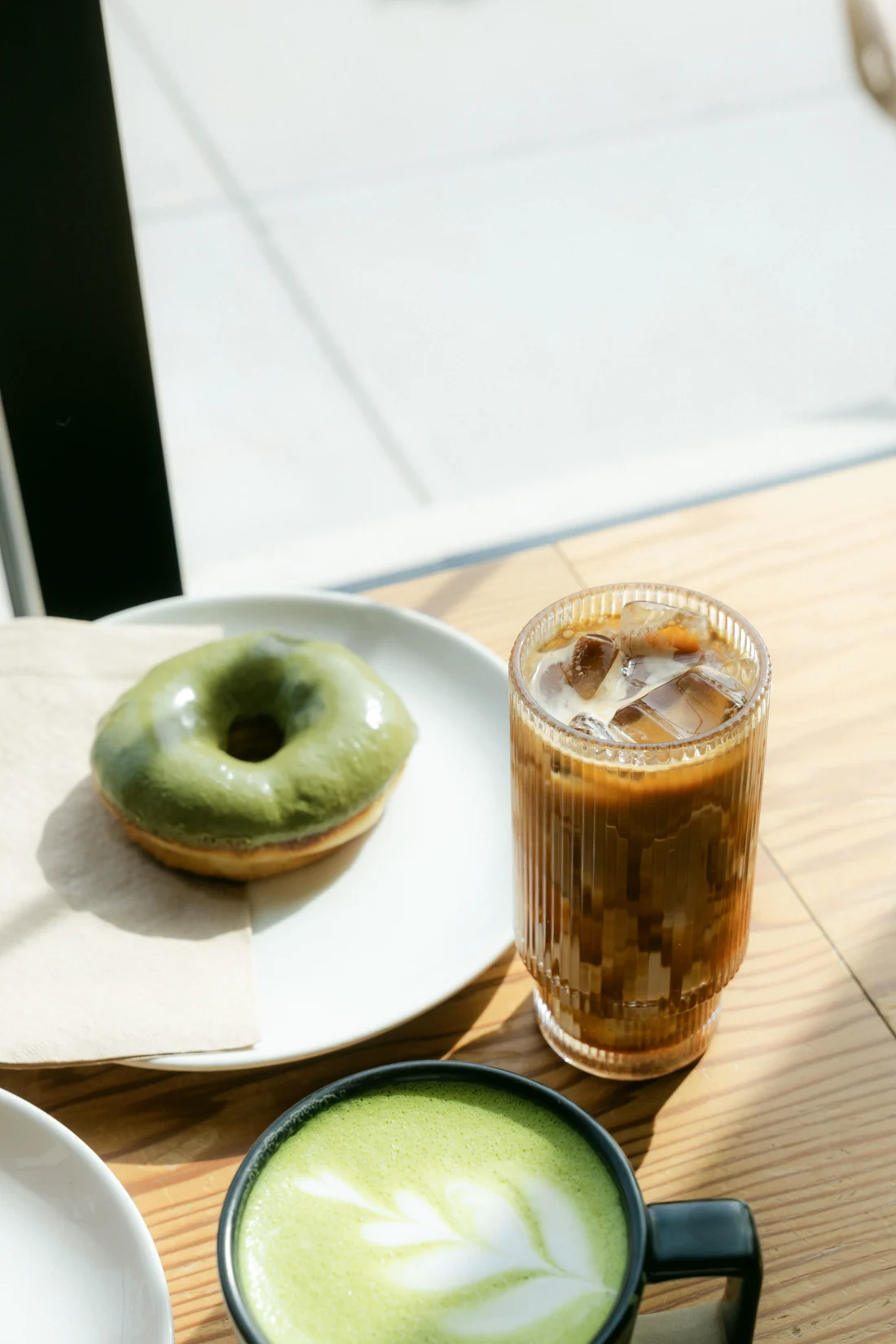 A matcha doughnut, an iced coffee and a matcha latte on a wooden café table from above